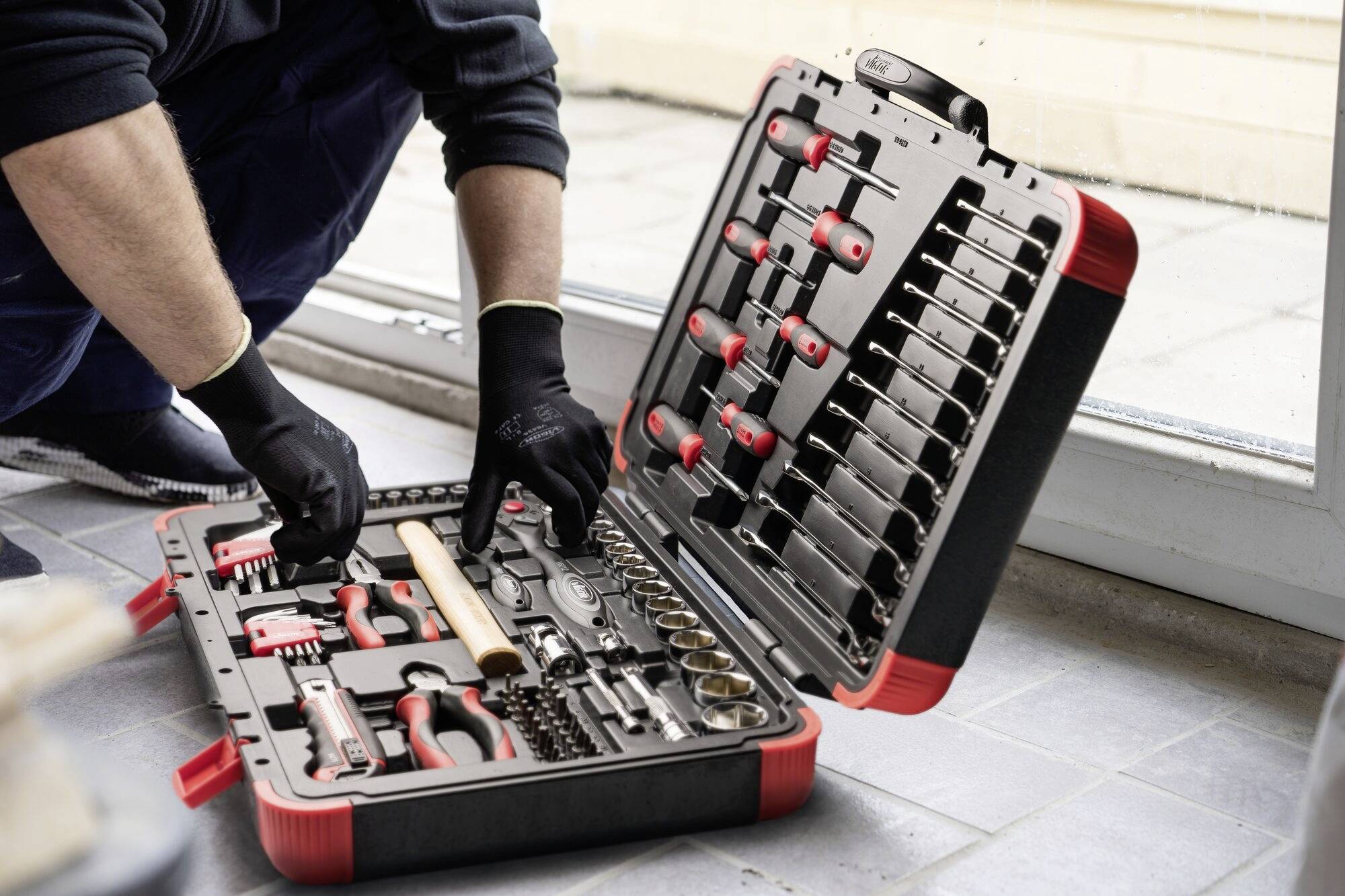 Eine Person in Handschuhen öffnet einen Werkzeugkoffer voller verschiedenartiger Werkzeuge, auf dem Boden liegend.