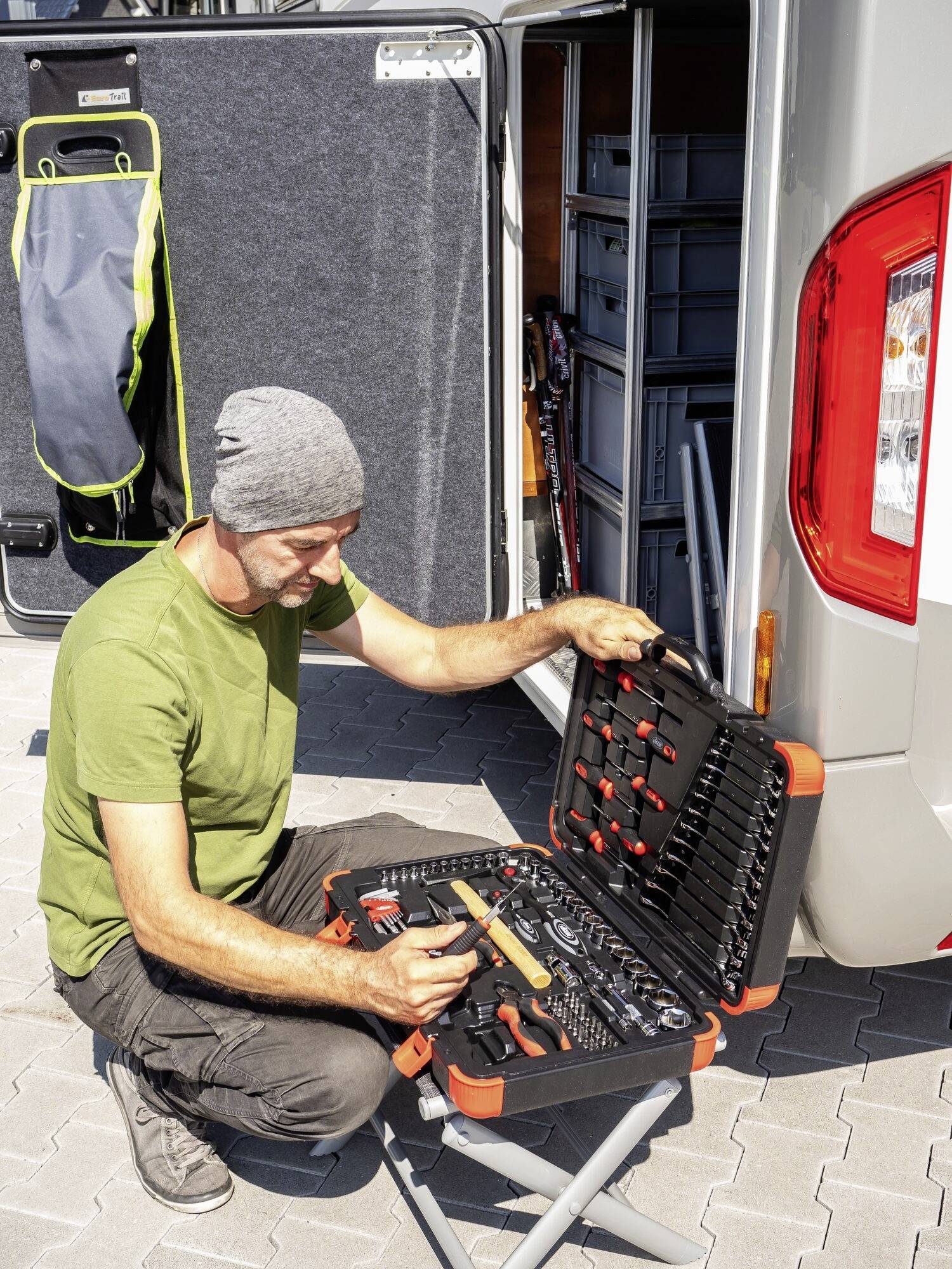 Ein Mann in einem grünen T-Shirt und grauer Mütze überprüft ein Werkzeugset vor einem offenen Wohnmobil.