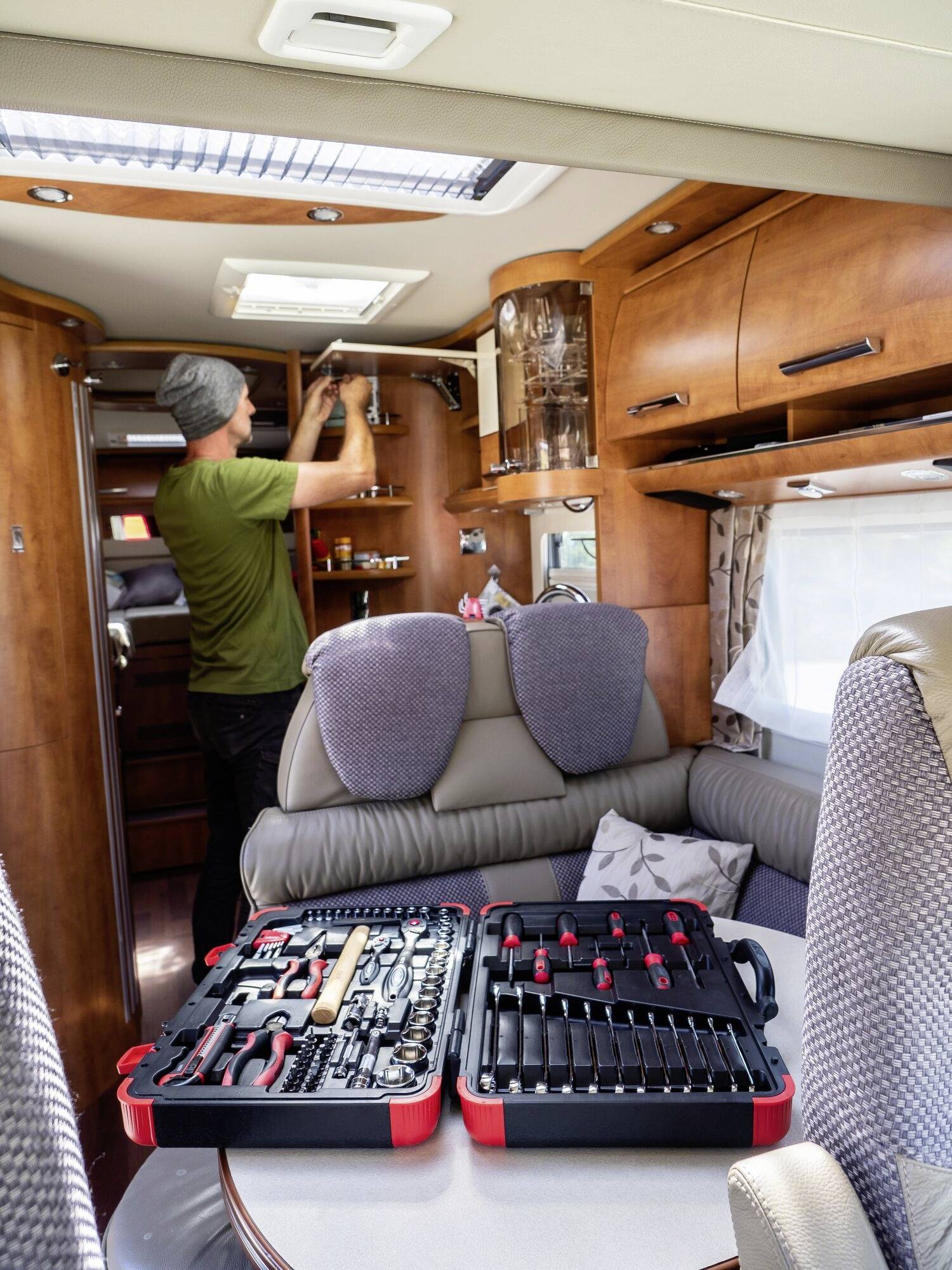 Ein Mann repariert ein Wohnmobil. Auf dem Tisch liegt ein offener Werkzeugkoffer. Der Innenraum wirkt wohnlich mit Holzmöbeln.