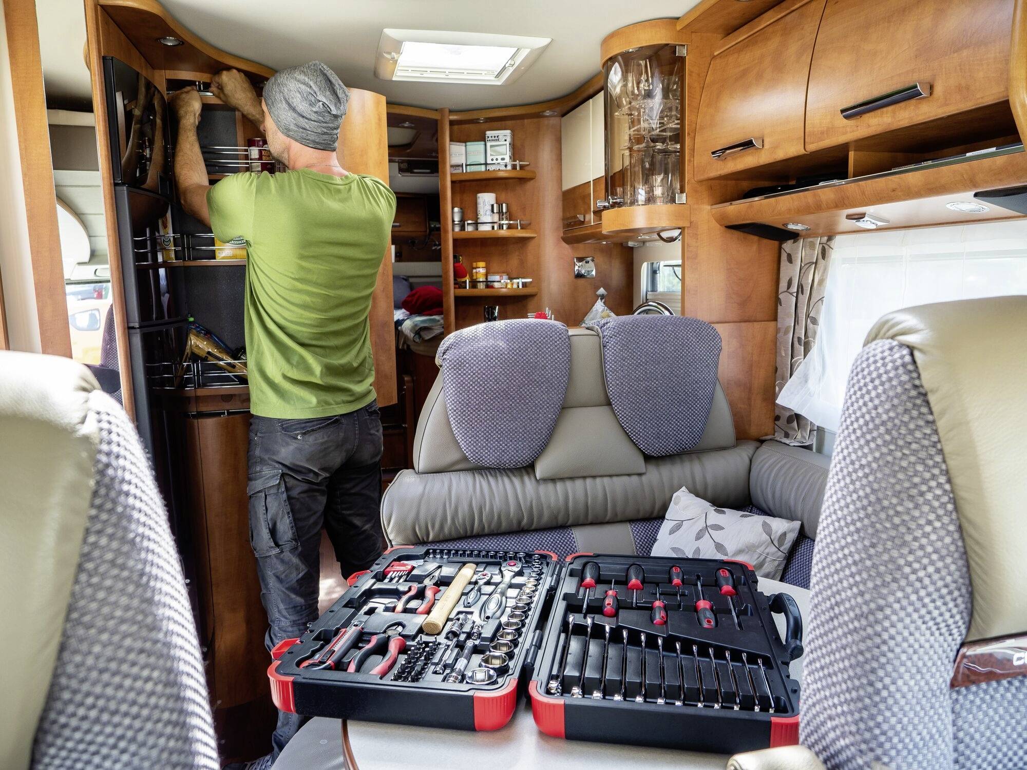Ein Mann repariert ein Schrank in einem Wohnmobil. Im Vordergrund steht ein geöffnetes Werkzeugset auf einem Tisch.