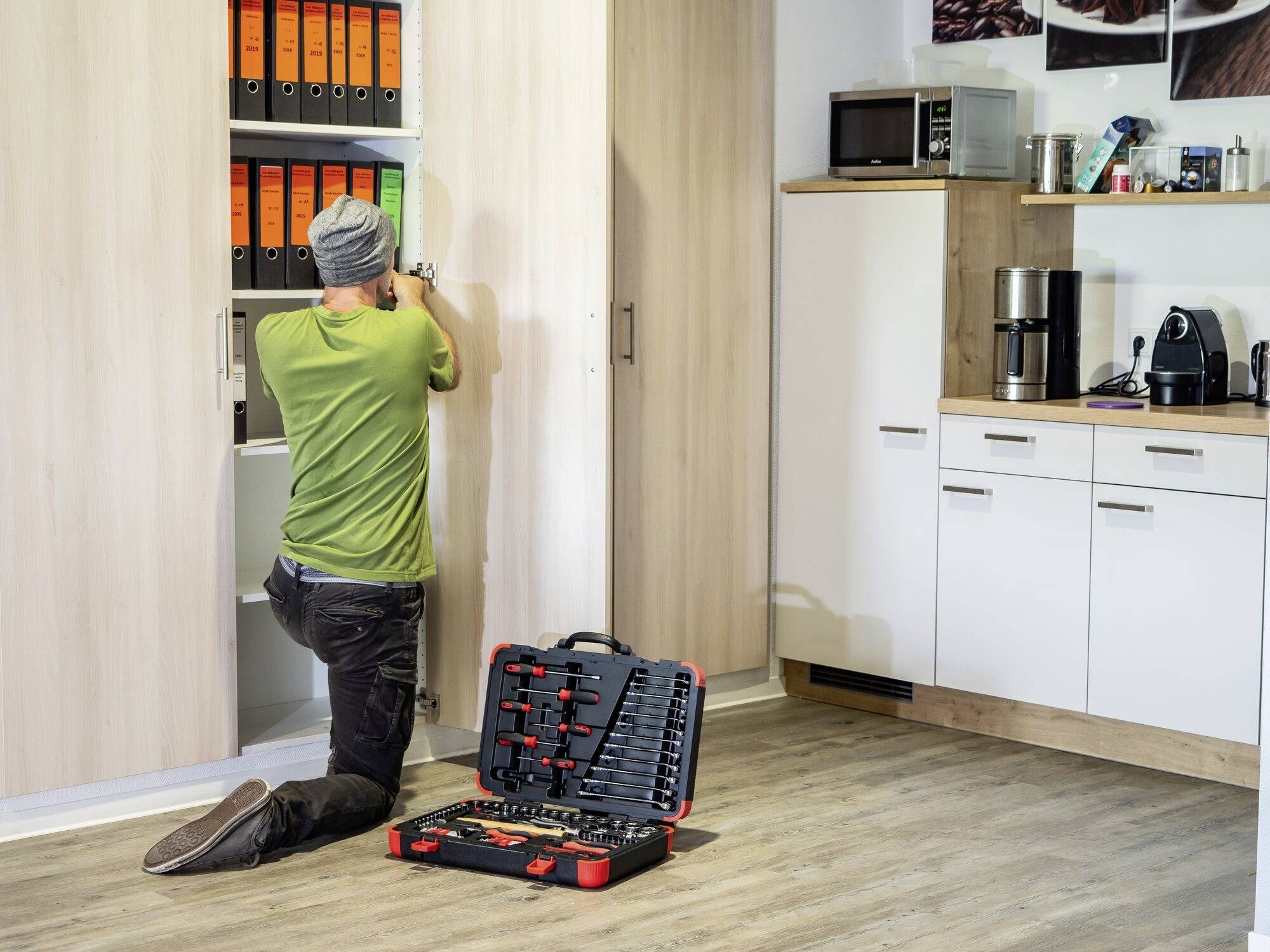 Eine Person in grüner Kleidung kniet vor einem Schrank in einer Küche und führt Reparaturen durch. Ein geöffneter Werkzeugkoffer liegt auf dem Boden.