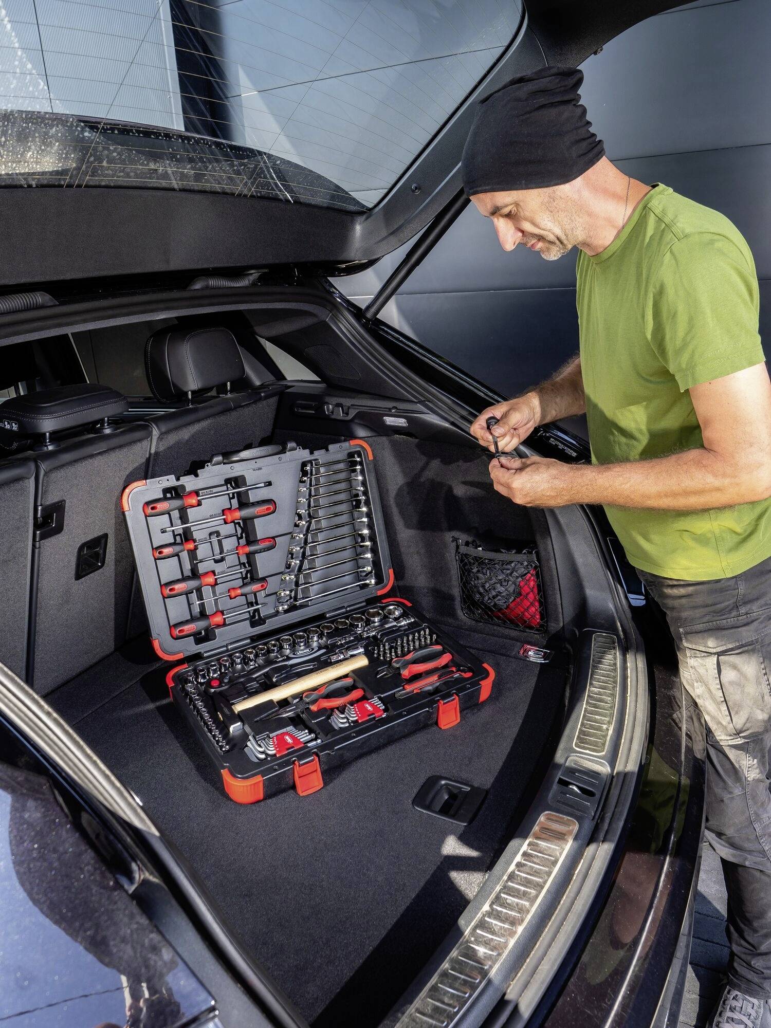 Ein Mann in grüner Kleidung steht am geöffneten Kofferraum eines Autos und überprüft eine Werkzeugkiste mit verschiedenen Werkzeugen.