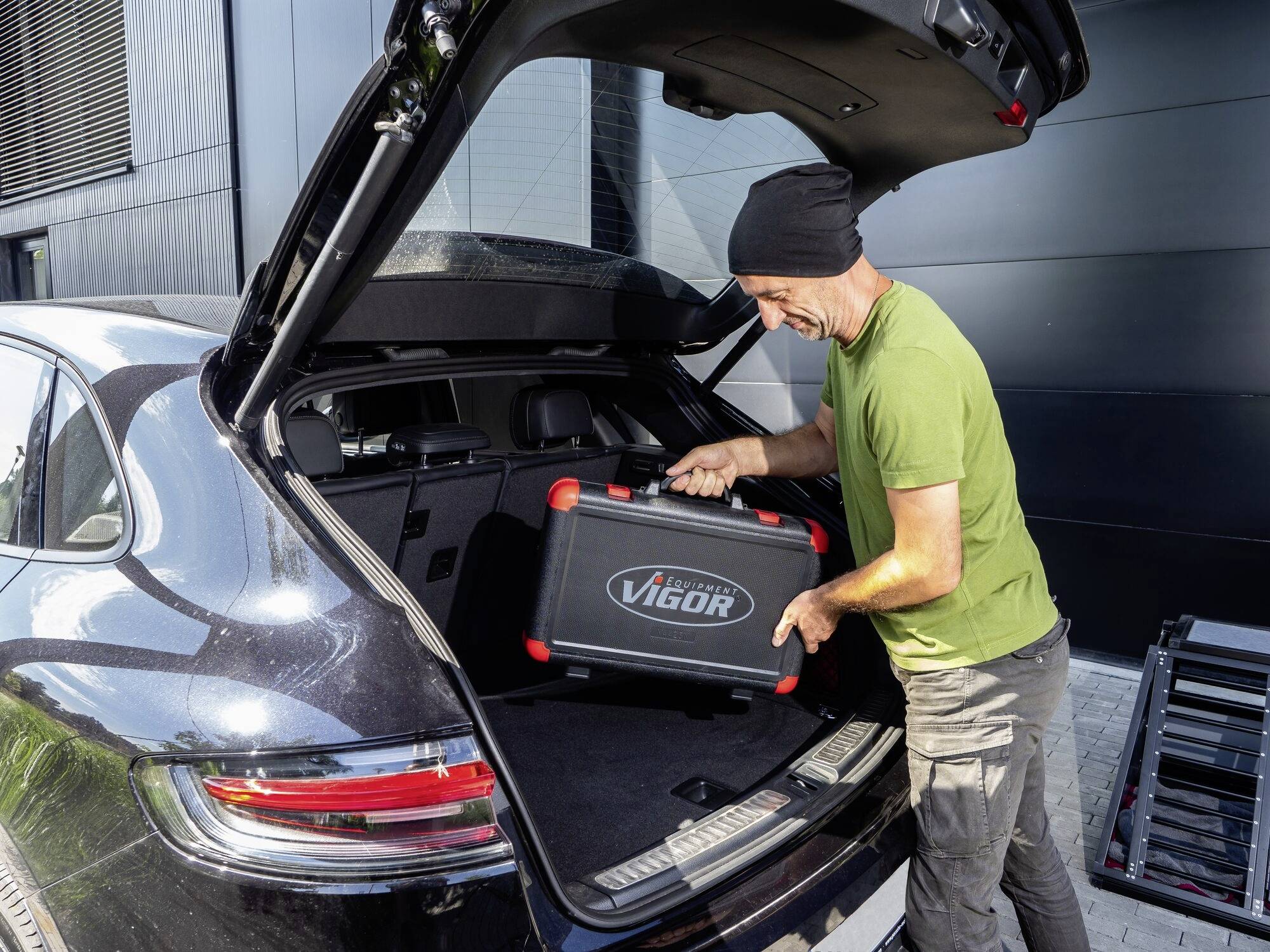 Ein Mann hebt einen Werkzeugkoffer aus dem Kofferraum eines dunklen Autos. Er trägt ein grünes T-Shirt und eine schwarze Mütze.