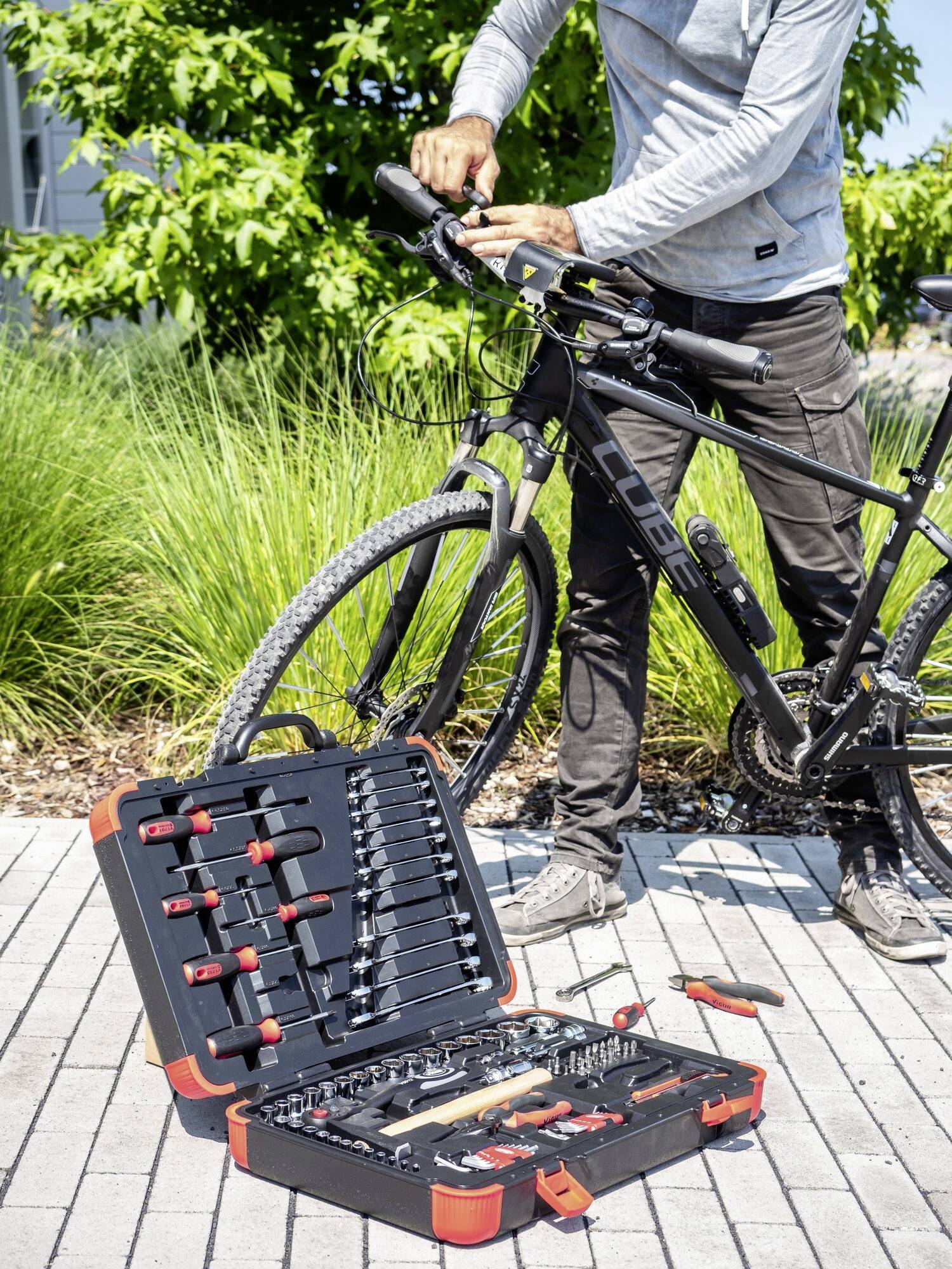 Ein Mann repariert ein Fahrrad im Freien, ein geöffneter Werkzeugkoffer mit verschiedenen Werkzeugen liegt auf dem Boden daneben.