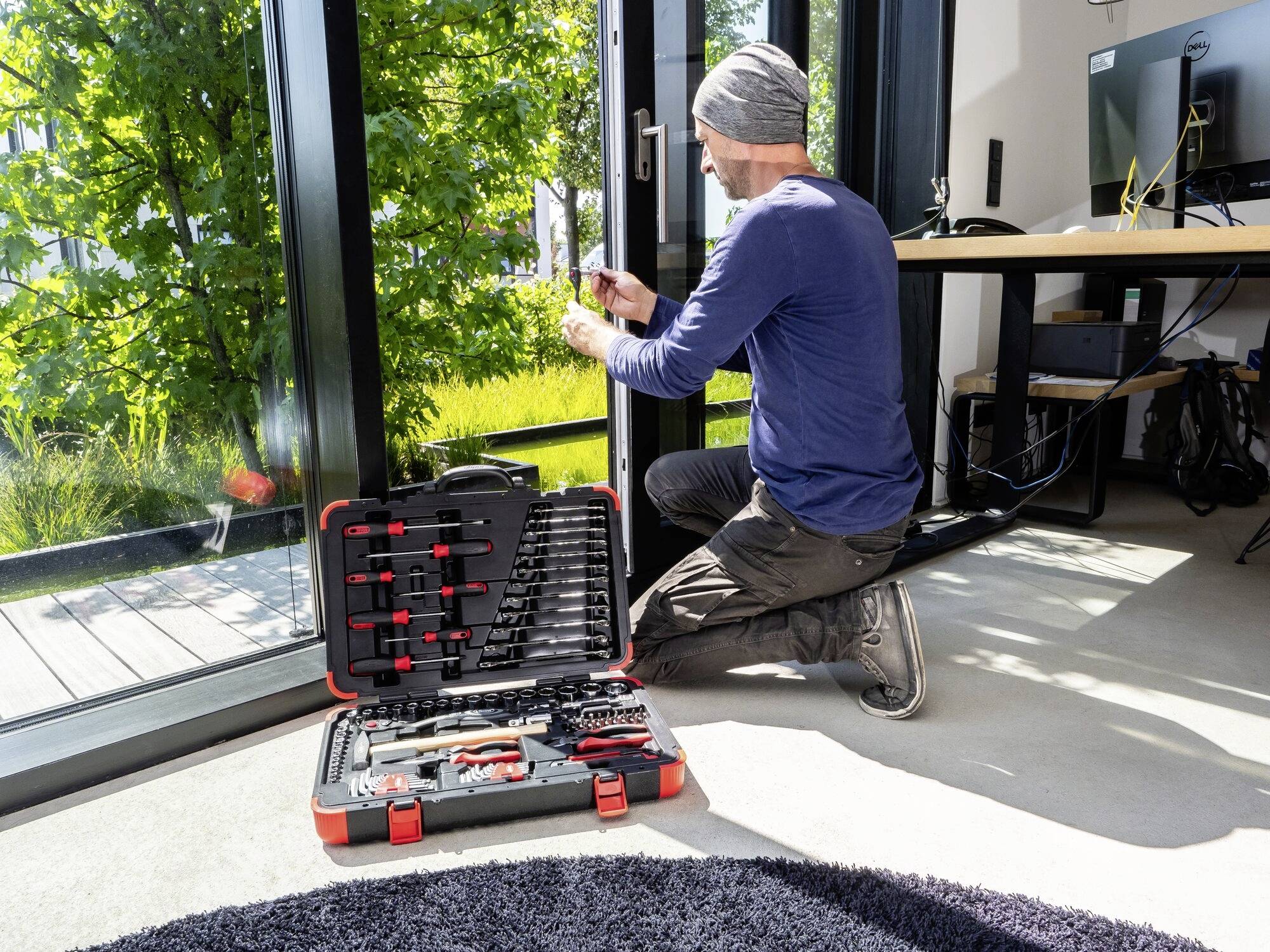 Ein Mann repariert eine Glastür mit einem Schraubenzieher. Ein offener Werkzeugkoffer liegt in der Sonne auf dem Boden.