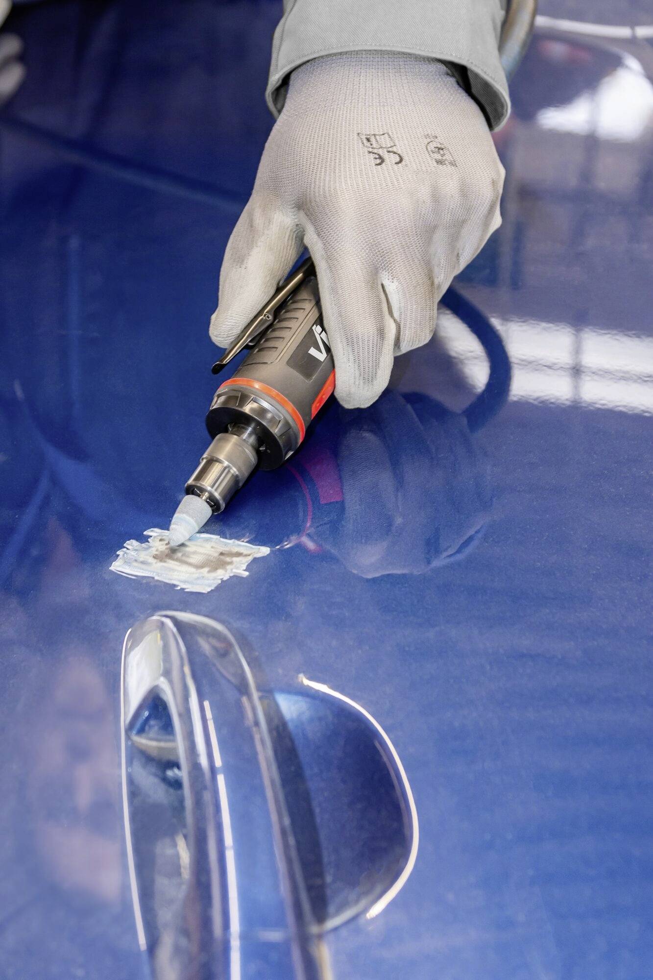 Eine Hand mit Handschuhen benutzt ein Elektrowerkzeug, um eine kleine beschädigte Stelle auf einer blauen Autotür zu reparieren.