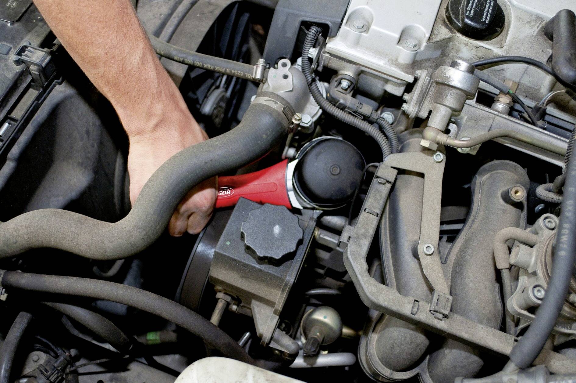 Eine Hand benutzt ein Werkzeug, um an einem Automotor zu arbeiten. Sichtbar sind verschiedene Motorenteile und Schläuche.