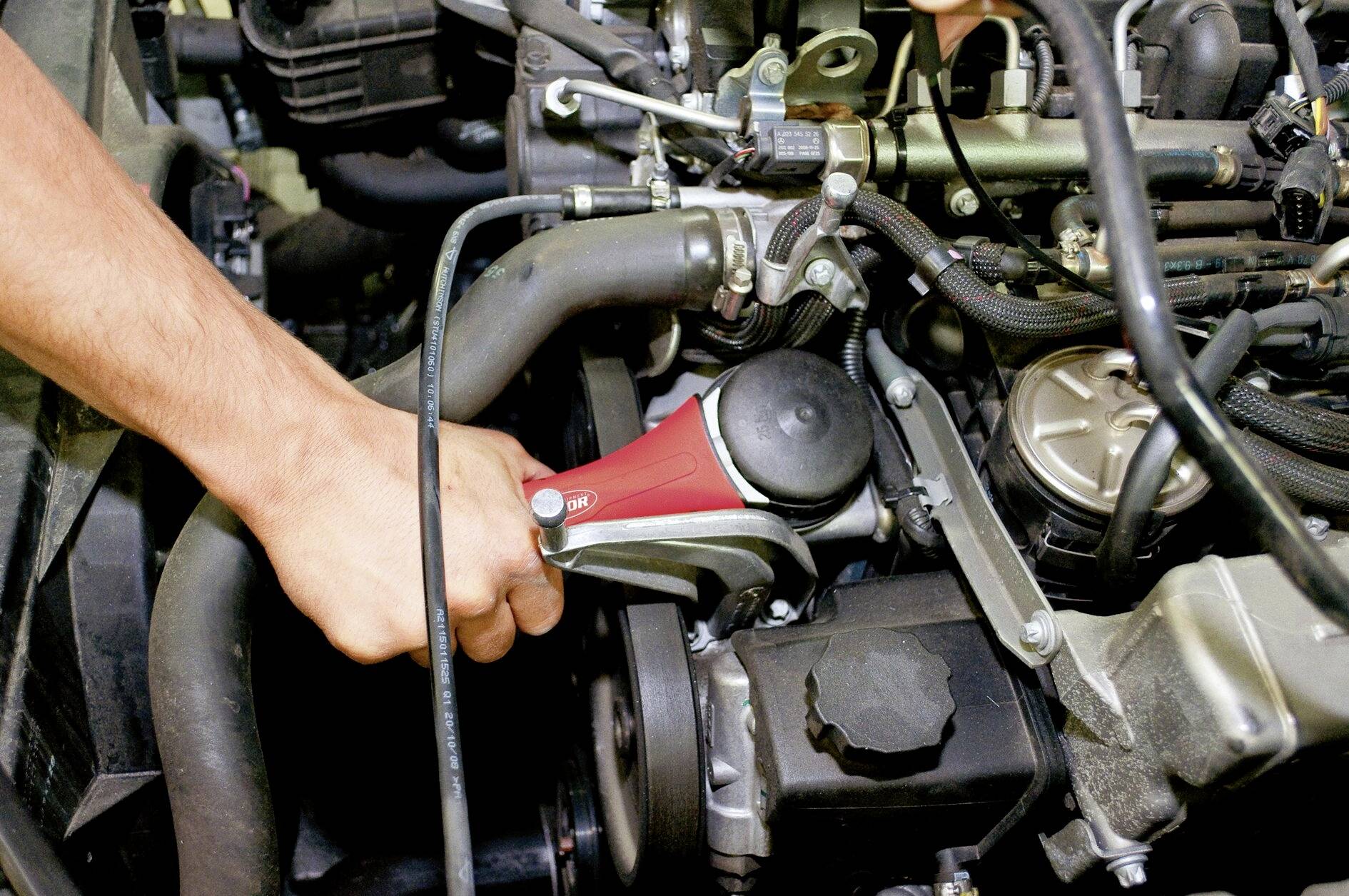 Eine Hand hält ein Werkzeug, das an einem Fahrzeugmotor arbeitet, umgeben von Schläuchen und mechanischen Teilen.