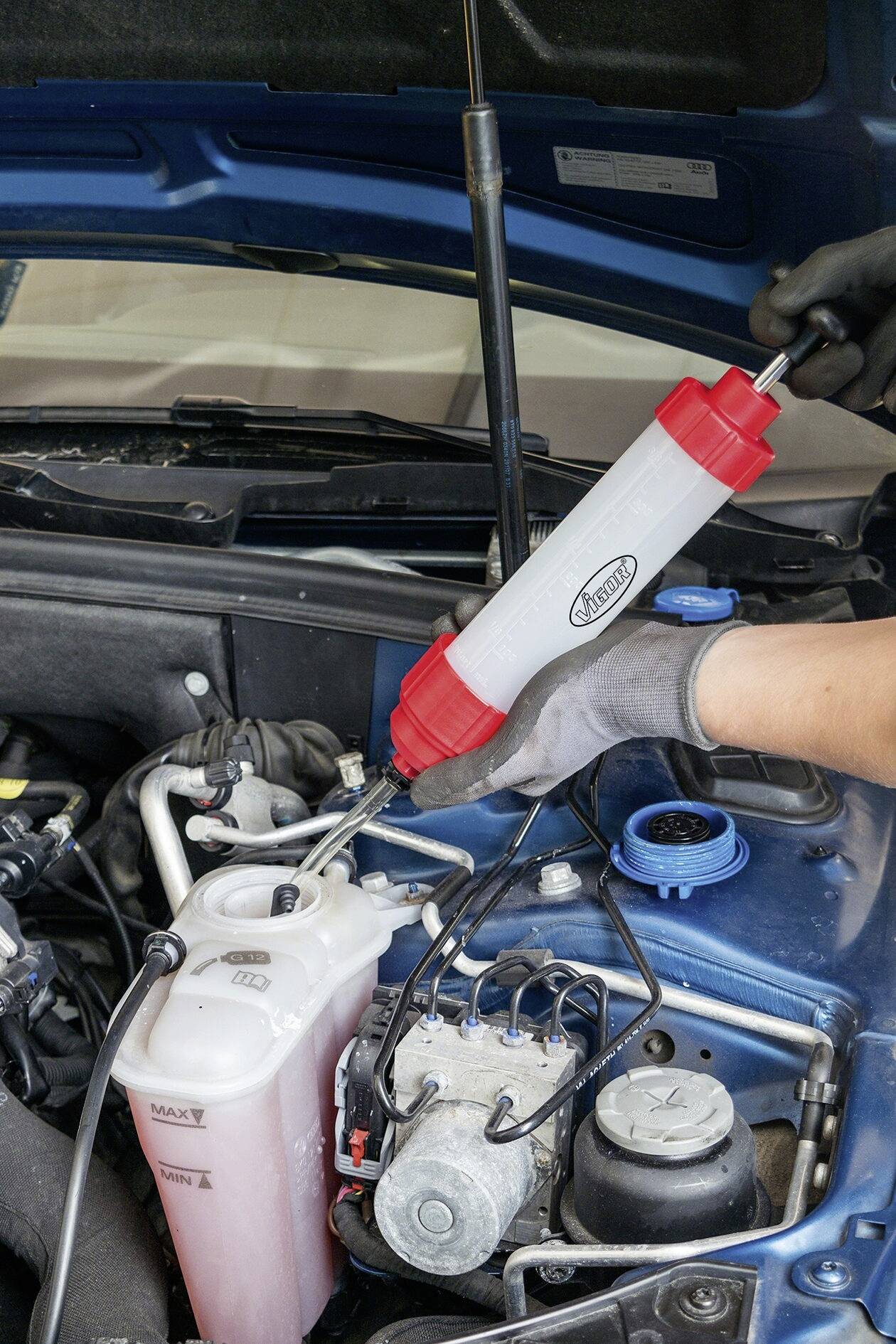 Eine Person füllt eine rote Flüssigkeit in einen weißen Behälter im Auto. Der Motorraum ist zu sehen, die Motorhaube ist offen.