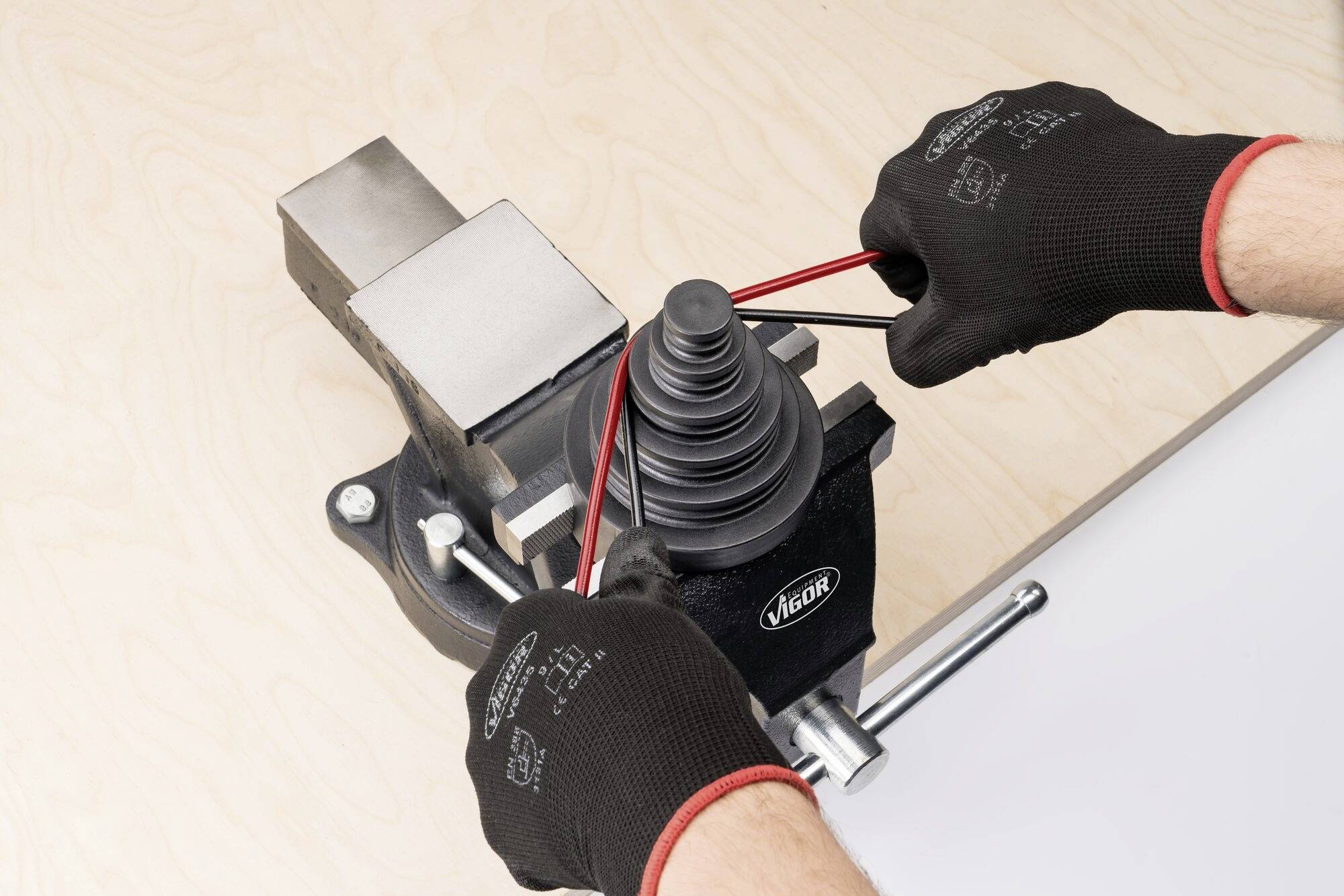 Zwei Hände in schwarzen Handschuhen befestigen einen roten Gummiring an einem Schraubstock mit einem federnden Mechanismus.