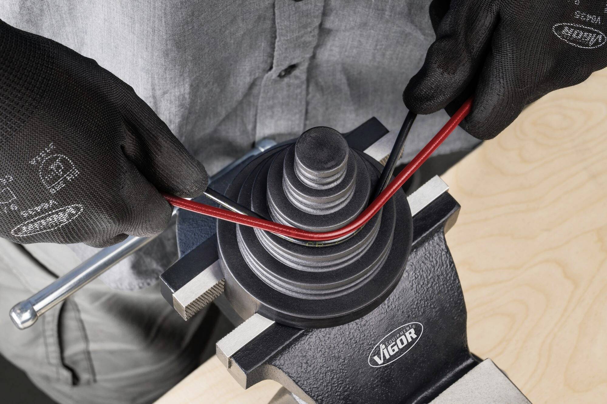 Eine Person biegt mit einem Rohrbiegegerät ein rotes Rohr. Das Gerät ist auf einem Tisch montiert. Die Person trägt schwarze Handschuhe.