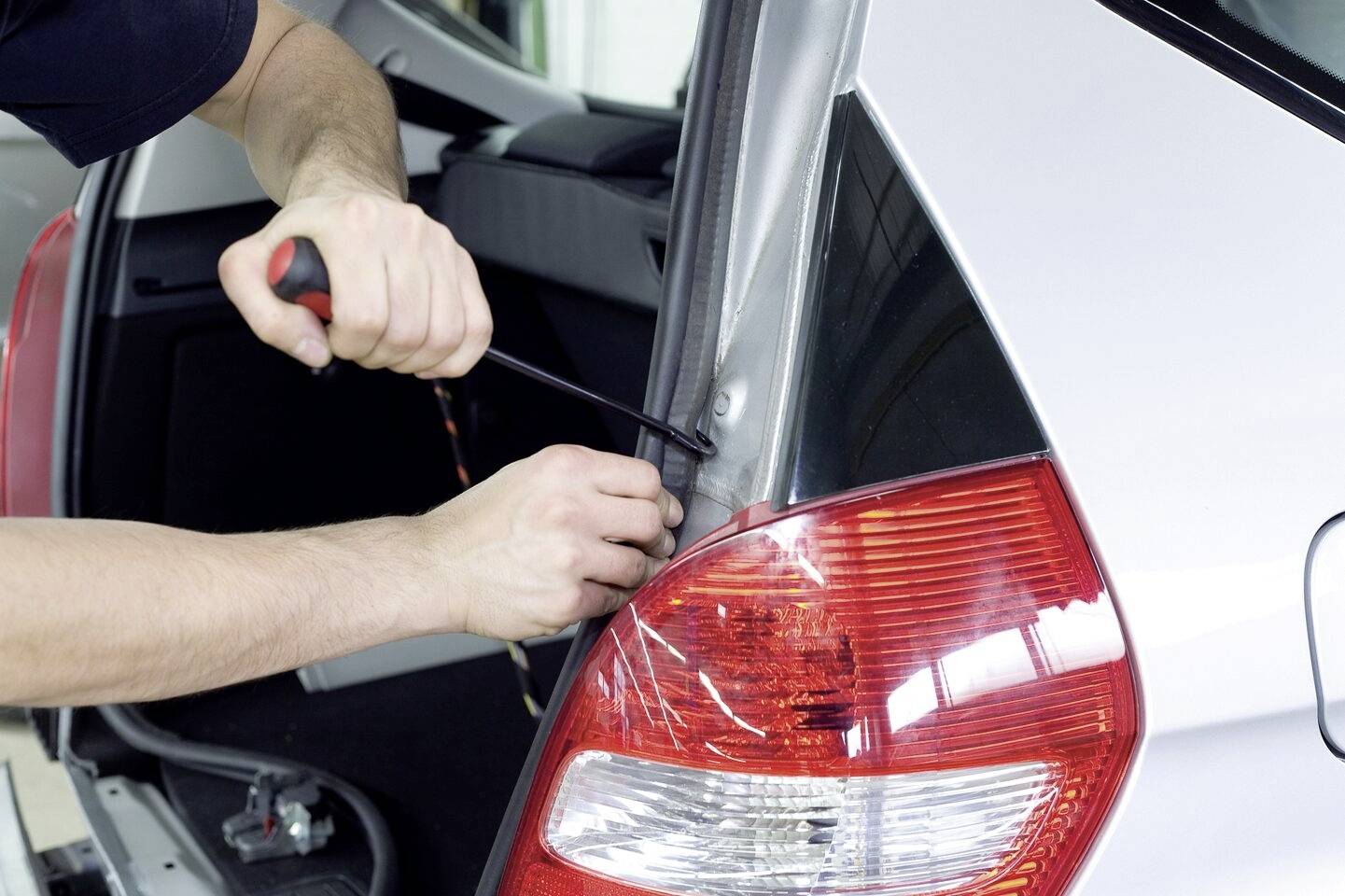 Eine Person repariert den hinteren linken Rücklichtbereich eines silbernen Autos mit einem Schraubenzieher.