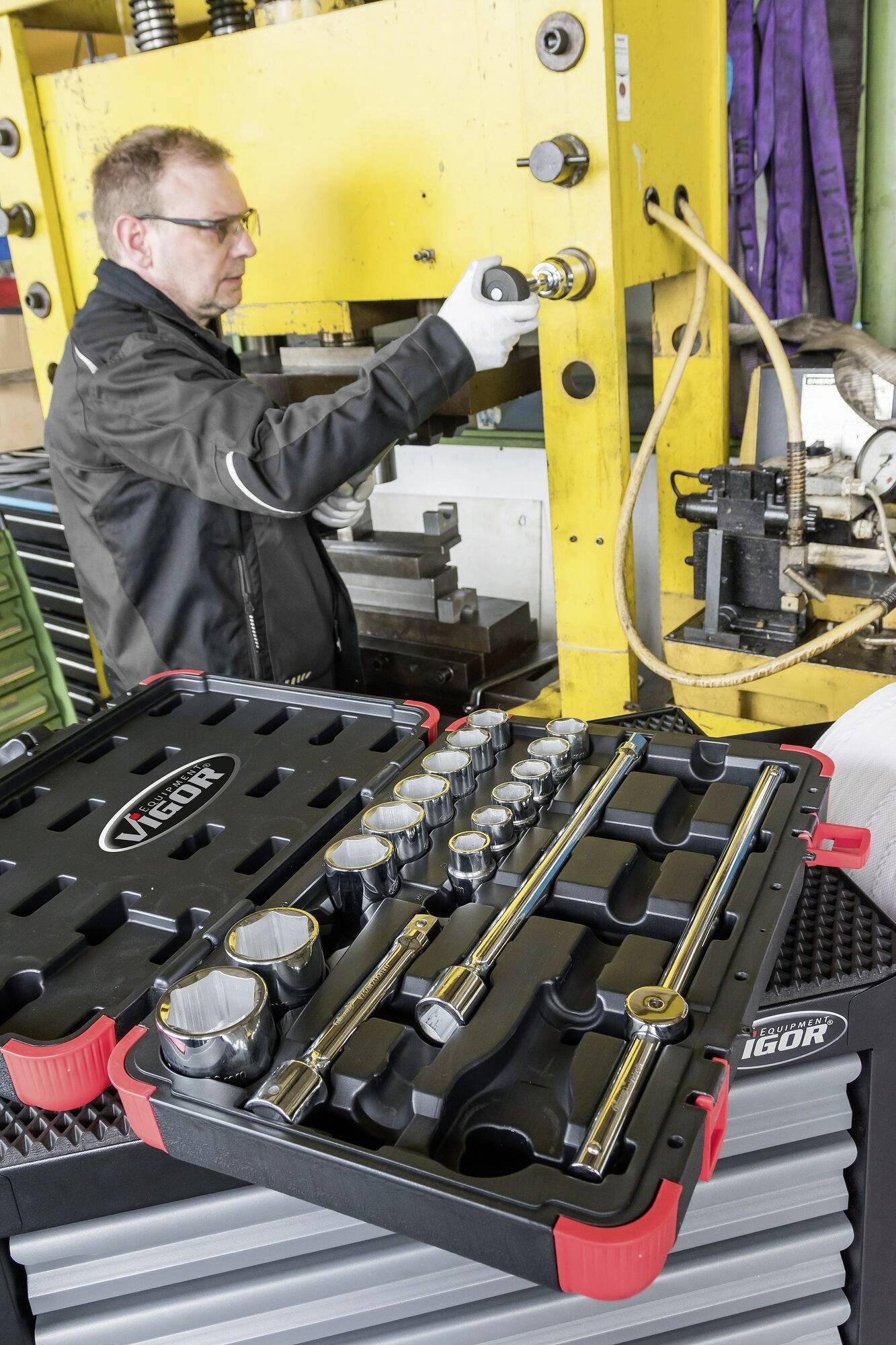 Eine Person arbeitet an einer Maschine in einer Fabrik. Im Vordergrund ist ein Werkzeugkasten mit verschiedenen Schraubenschlüsseln und Steckaufsätzen zu sehen.