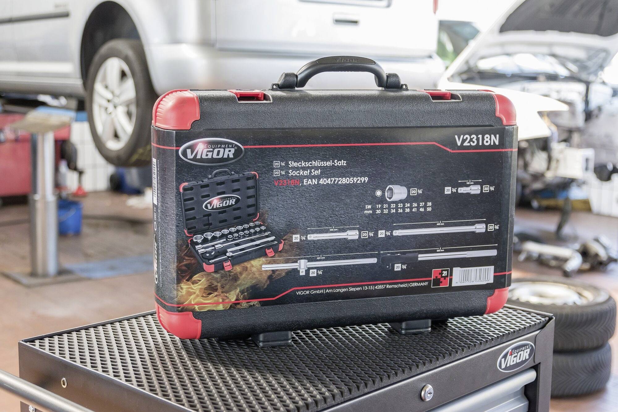 Ein Werkzeugkoffer von Vigor, Modell V2318N, steht vor einem Auto in einer Werkstatt. Aufgedruckte Grafiken zeigen den Inhalt des Sets.