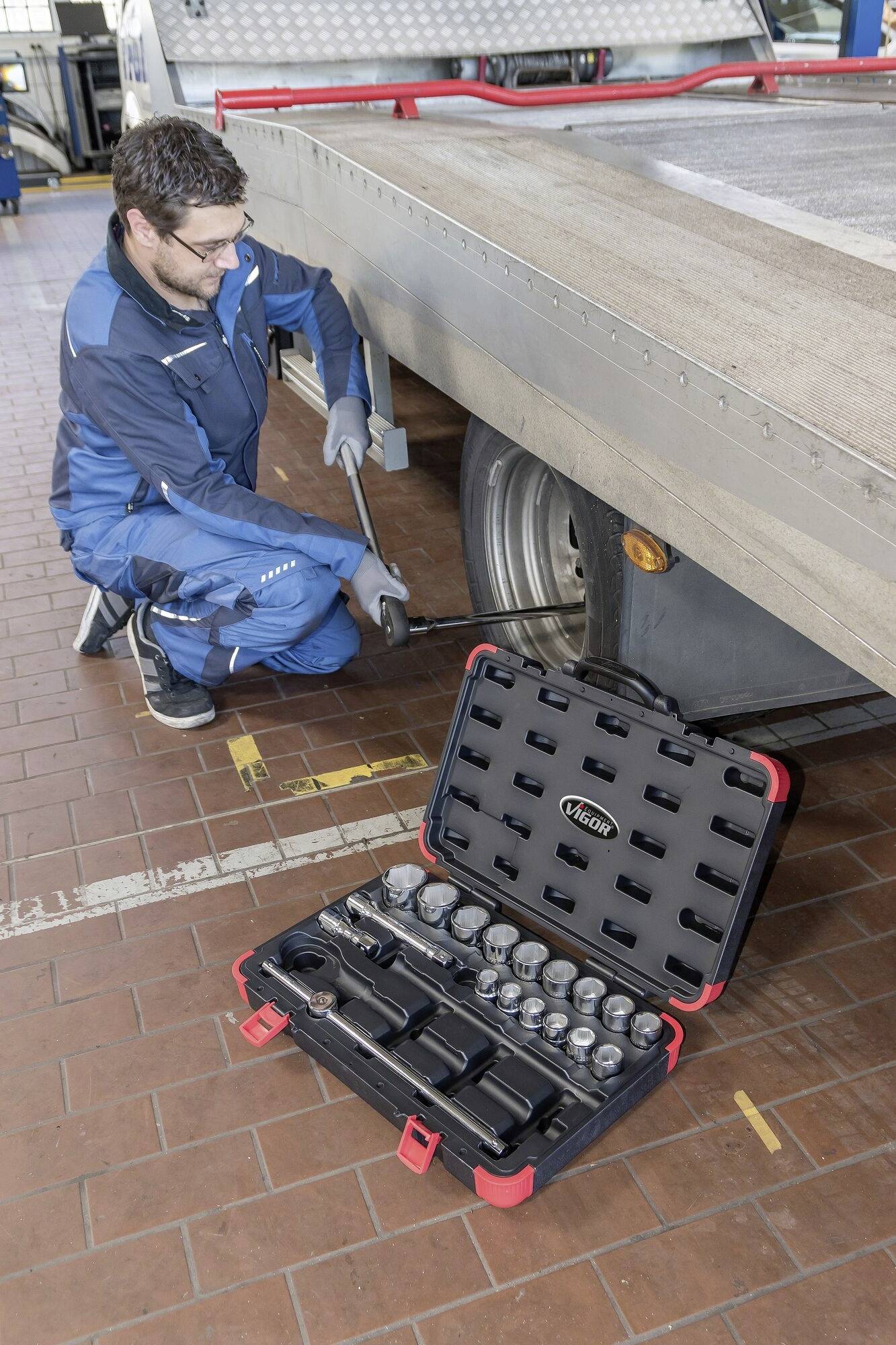 Ein Mechaniker in blauer Arbeitskleidung überprüft die Räder eines Anhängers mit einem Werkzeugkasten voller Schraubenschlüssel im Vordergrund.