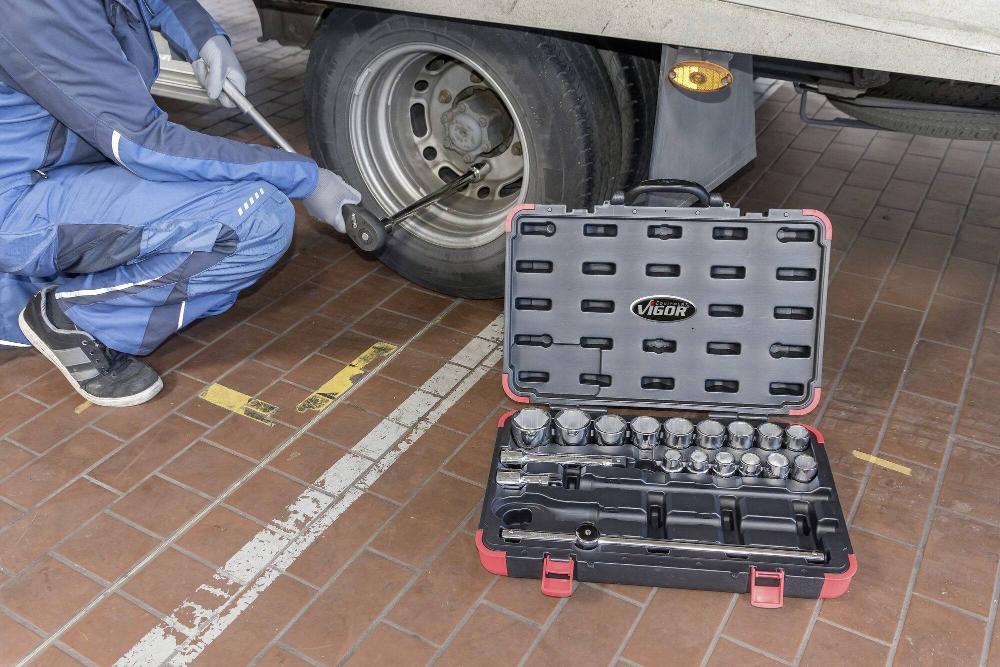 Eine Person wechselt ein Reifen eines Fahrzeugs. Im Vordergrund befindet sich ein geöffneter Werkzeugkoffer mit verschiedenen Aufsätzen.
