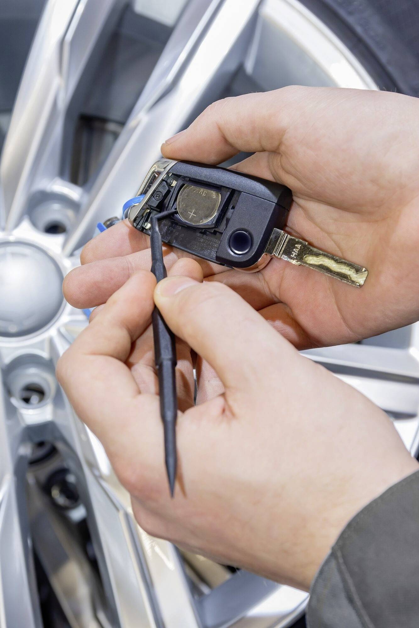 Eine Nahaufnahme zeigt Hände, die einen Autoschlüssel mit einem Werkzeug reparieren oder öffnen. Im Hintergrund ist ein Autorad sichtbar.