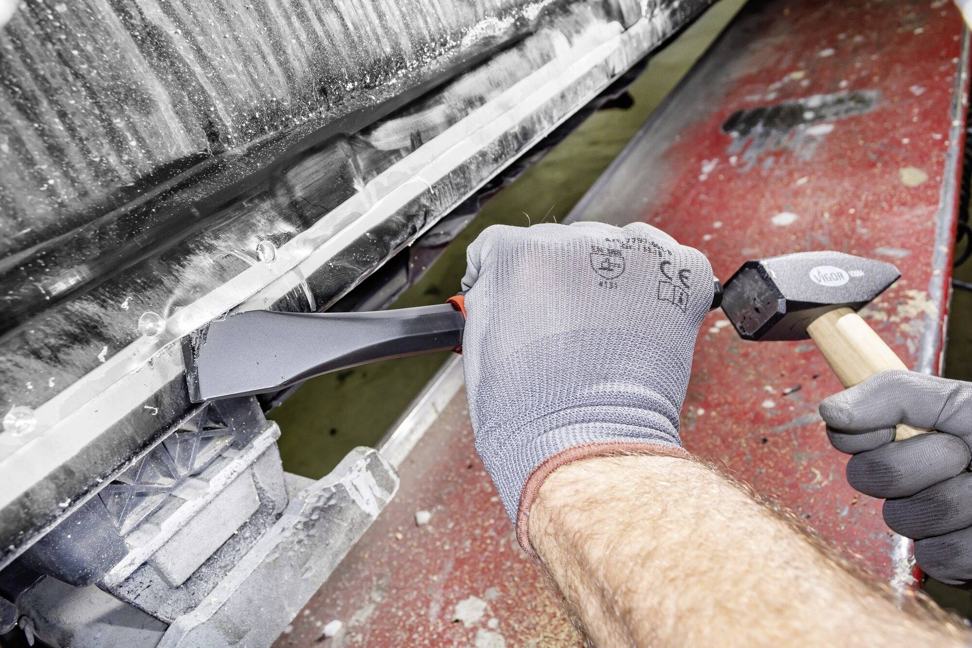 'Eine Person trägt Handschuhe und arbeitet mit einem Meißel und Hammer an einem Metallteil. Es sind Werkzeuge und Schmutz sichtbar.'