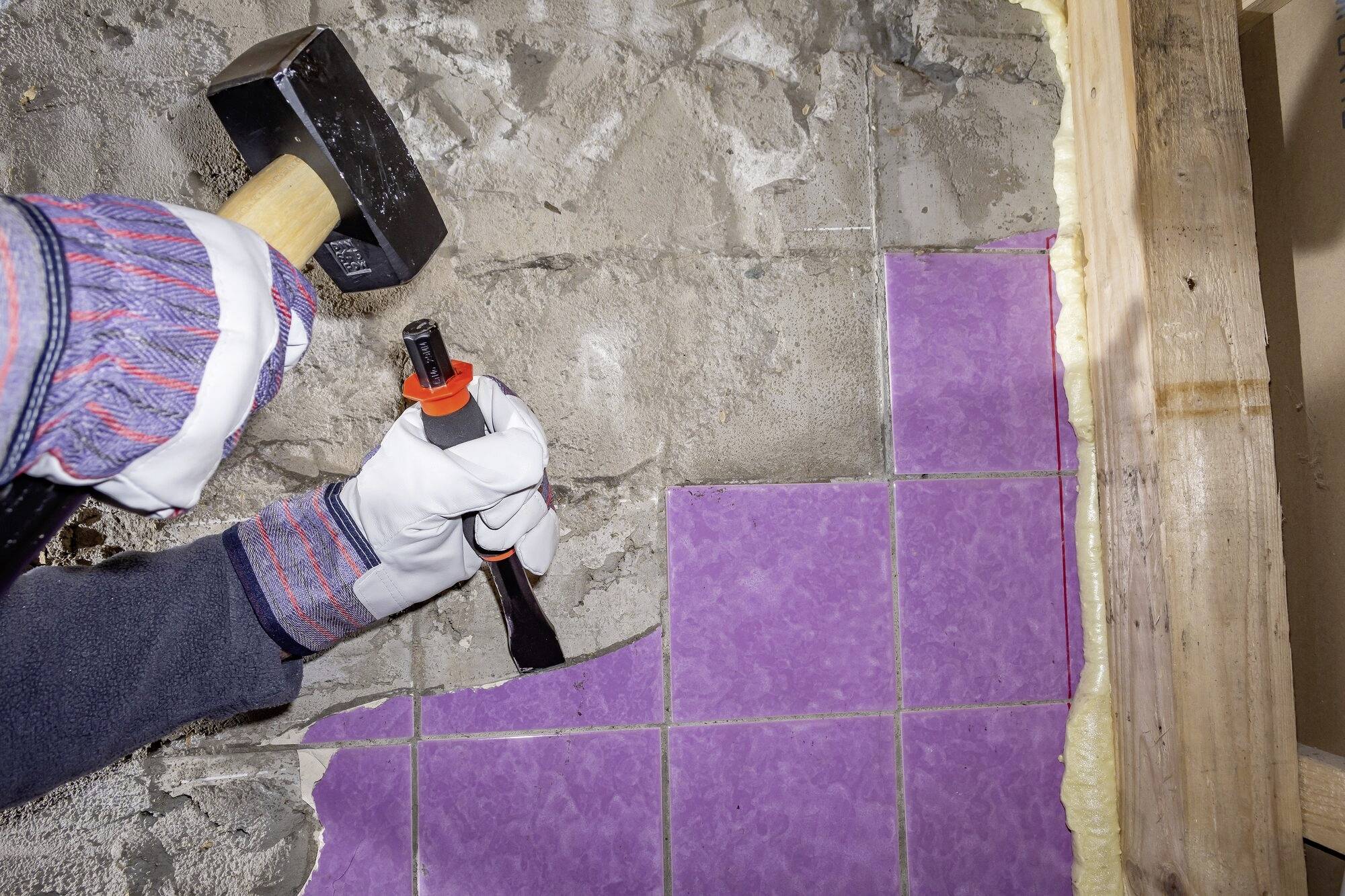 Person mit Hammer und Meißel entfernt lila Fliesen von einer Wand. Hand trägt Schutzhandschuhe. Holzrahmen am Rand sichtbar.