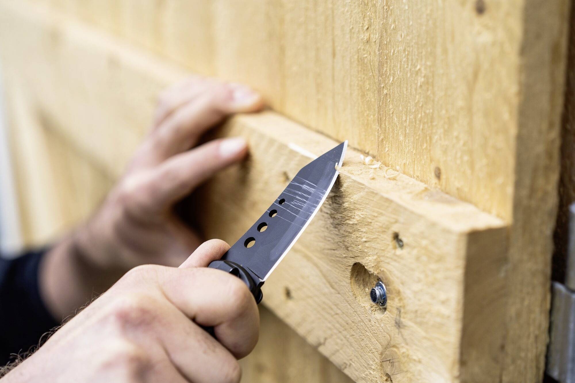 Eine Person schnitzt mit einem schwarzen Messer Holz, das an einer Wand befestigt ist.