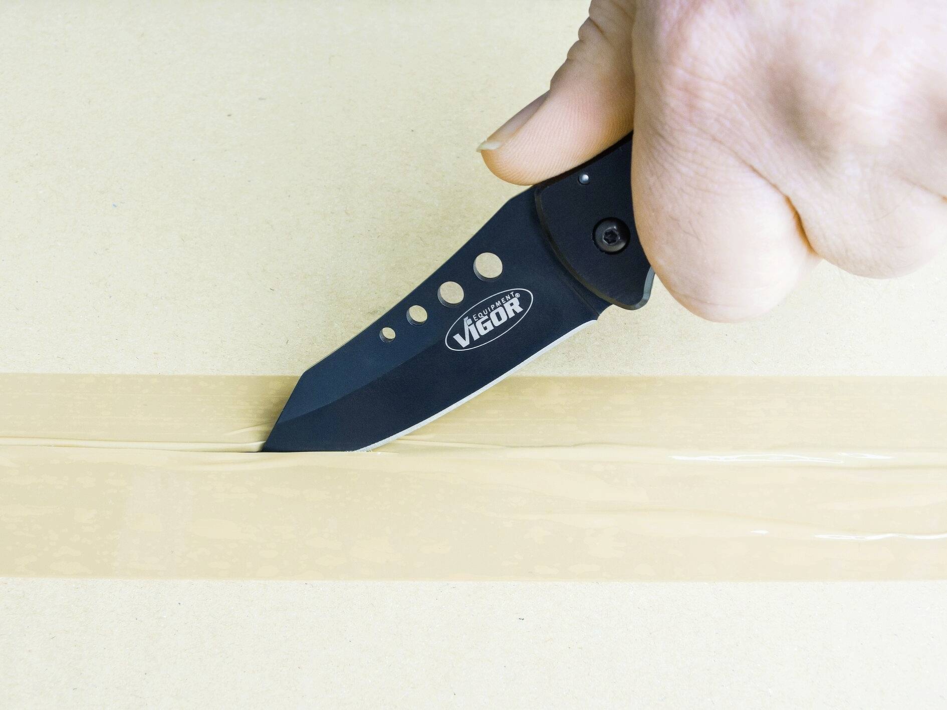 Eine Hand schneidet mit einem schwarzen Klappmesser einen Karton entlang eines Klebebandes auf.