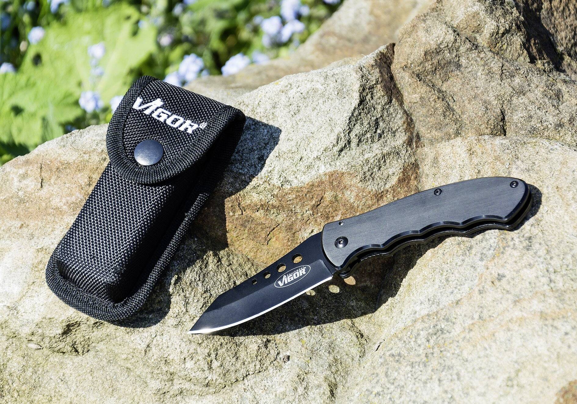 Ein Klappmesser mit schwarzem Griff liegt neben einer schwarzen Nylonhülle auf einem Felsen. Markenname 'Vigor' auf beiden Teilen sichtbar.