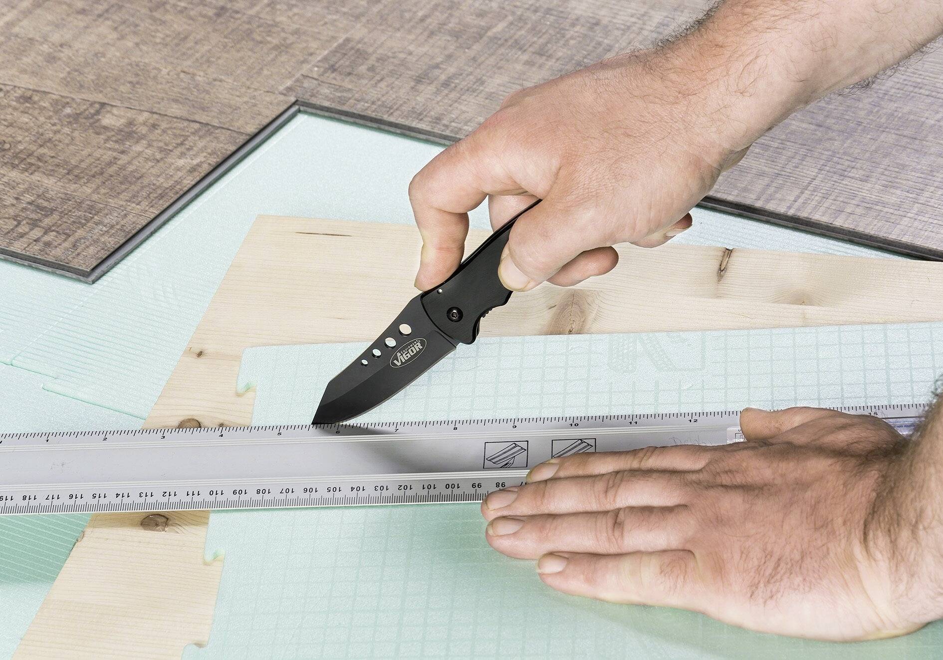 'Person schneidet mit einem schwarzen Messer eine transparente Kunststoffplatte entlang eines Lineals auf einem Holztisch.'