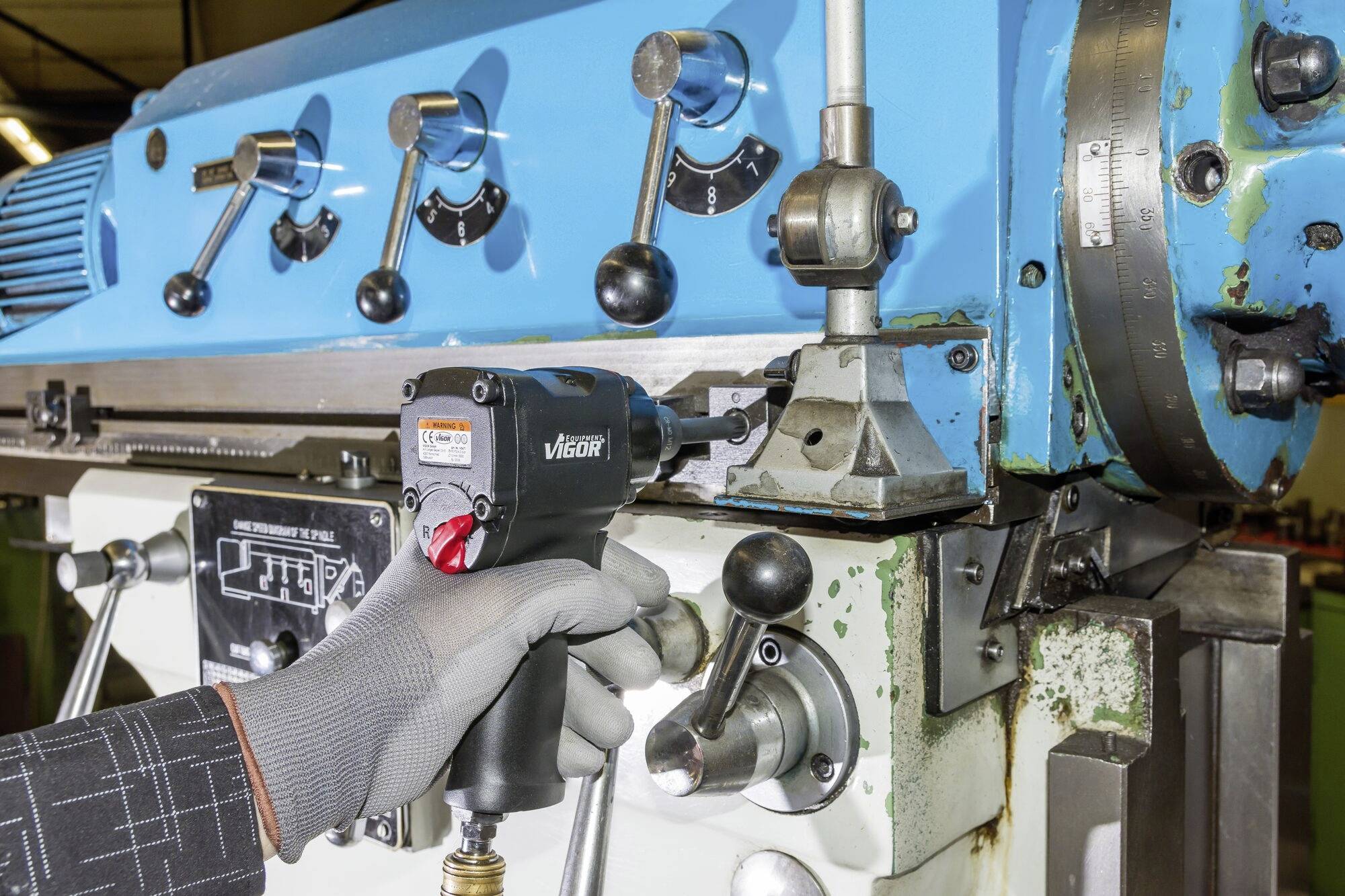 Eine Person bedient eine blaue Industriemaschine mit einem Elektrowerkzeug, um einen Mechanismus anzupassen. Verschiedene Hebel und Zifferblätter sind erkennbar.
