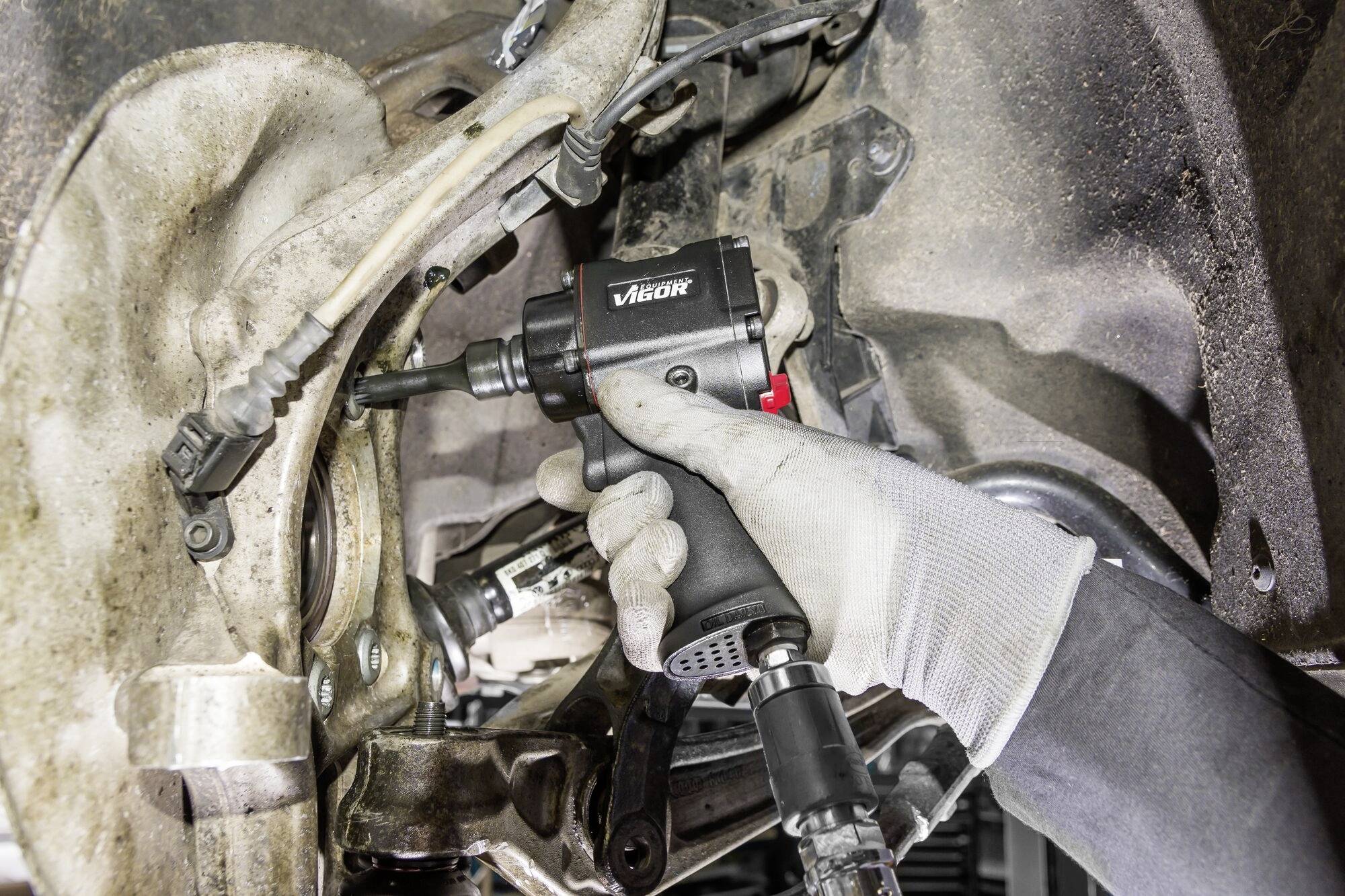 Mechanikerhand in Schutzhandschuh bedient eine Druckluft-Ratschenschlüssel, um eine Schraube an einer Fahrzeugachse zu fixieren.