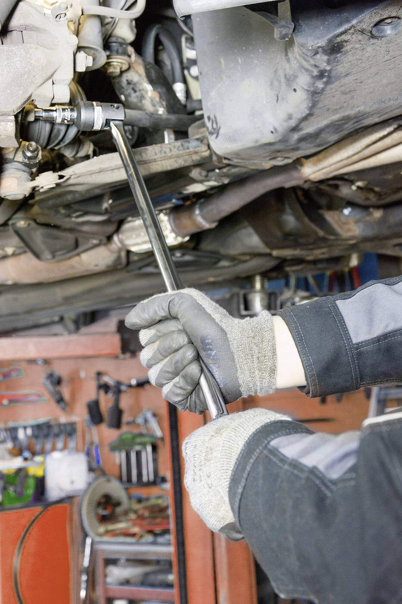 Eine Person in Arbeitskleidung repariert einen Auto-Unterboden in einer Werkstatt, hält ein Werkzeug an ein Autoteil.