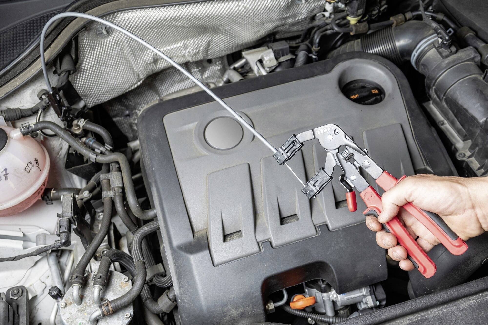 Eine Hand hält Zangen über einem Automotor, umgeben von Kabeln und Schläuchen. Fokus auf Wartungsarbeiten am Motor.