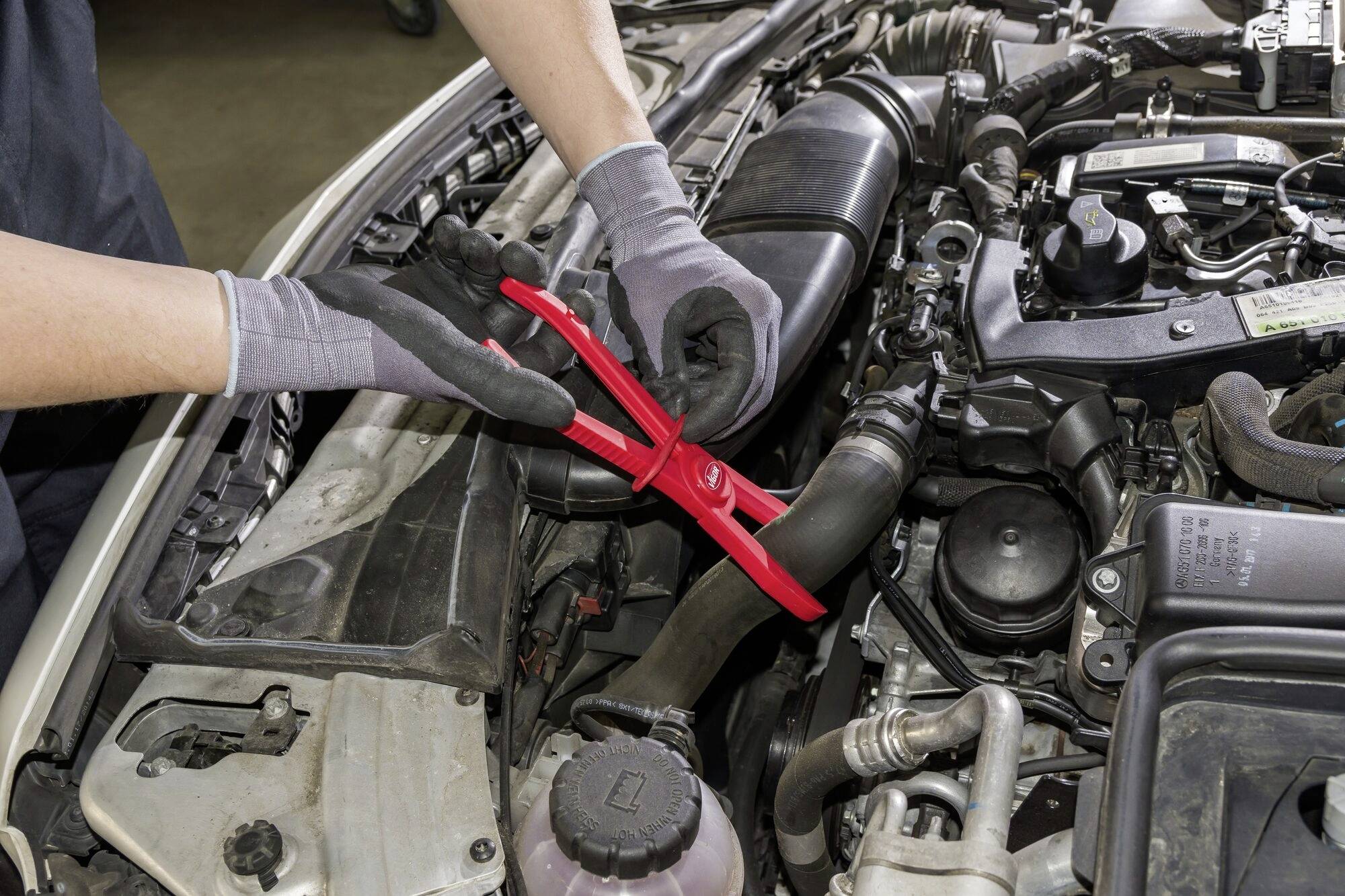 Mechaniker verwendet Zange, um Teil im Motorraum eines Autos zu reparieren. Hände tragen Arbeitshandschuhe.