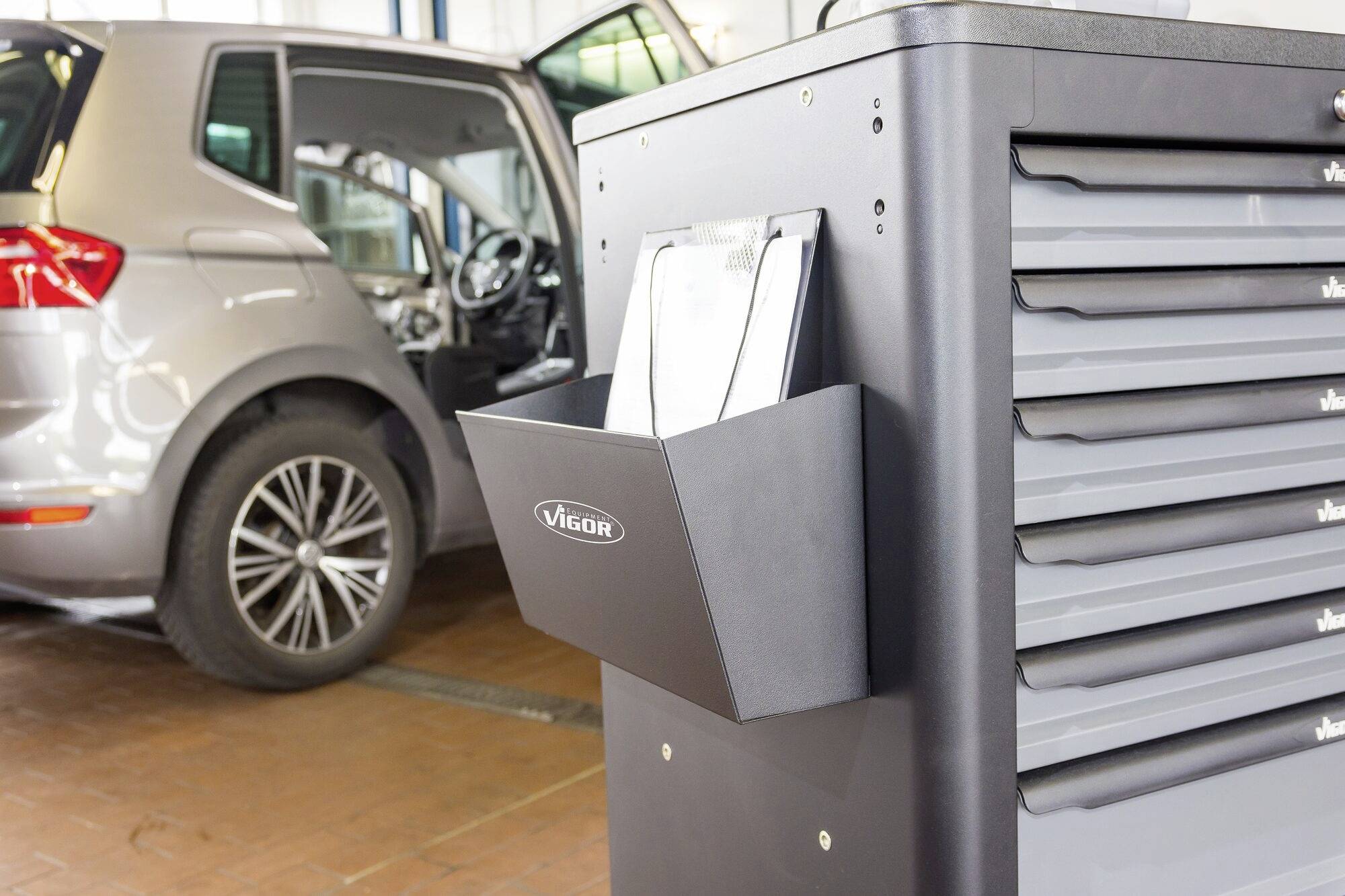 Ein graues Auto steht in einer Werkstatt. Ein geöffnetes Werkzeugschrank-Fach zeigt eine Klemmbretthalterung an der Seite.