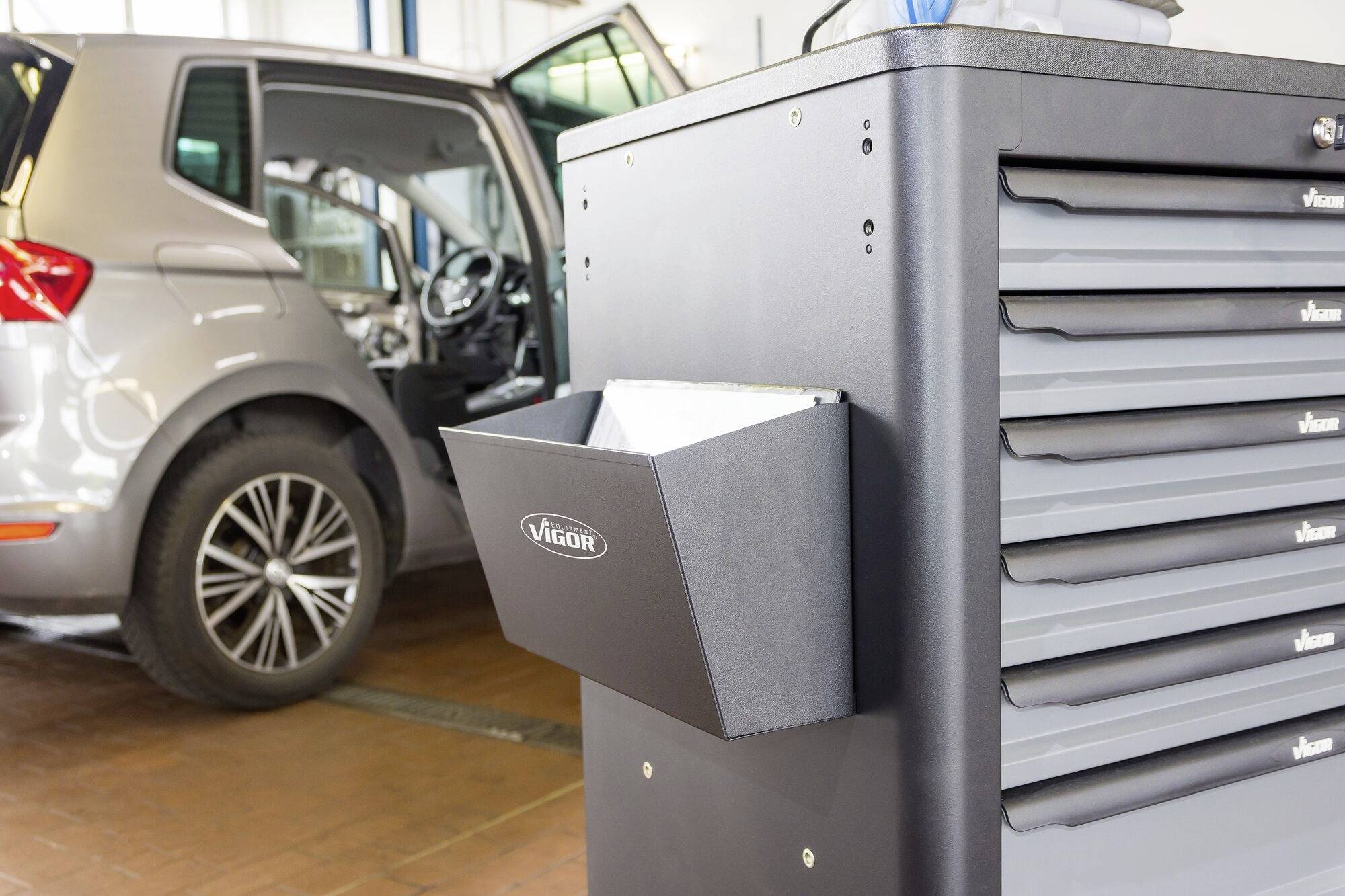 Ein graues Auto wird in einer Werkstatt repariert, nebendran steht eine schwarze Werkzeugschublade mit dem Schriftzug 'Vigor'.