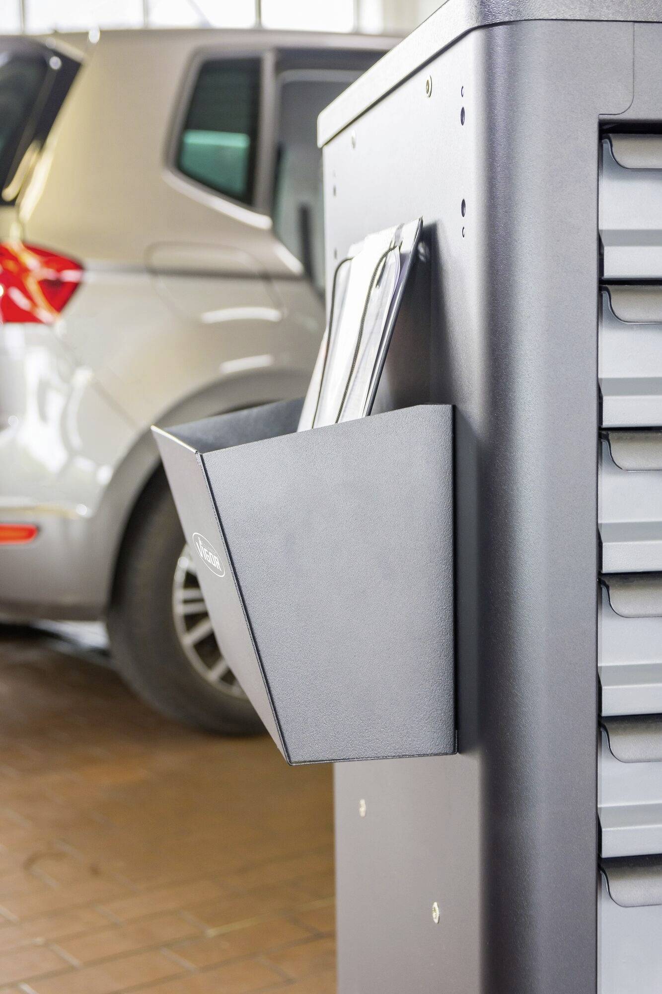 Ein Auto steht in einer Werkstatt. Daneben befindet sich ein Werkzeugschrank mit einem Fach, in dem Papiere oder Dokumente liegen.