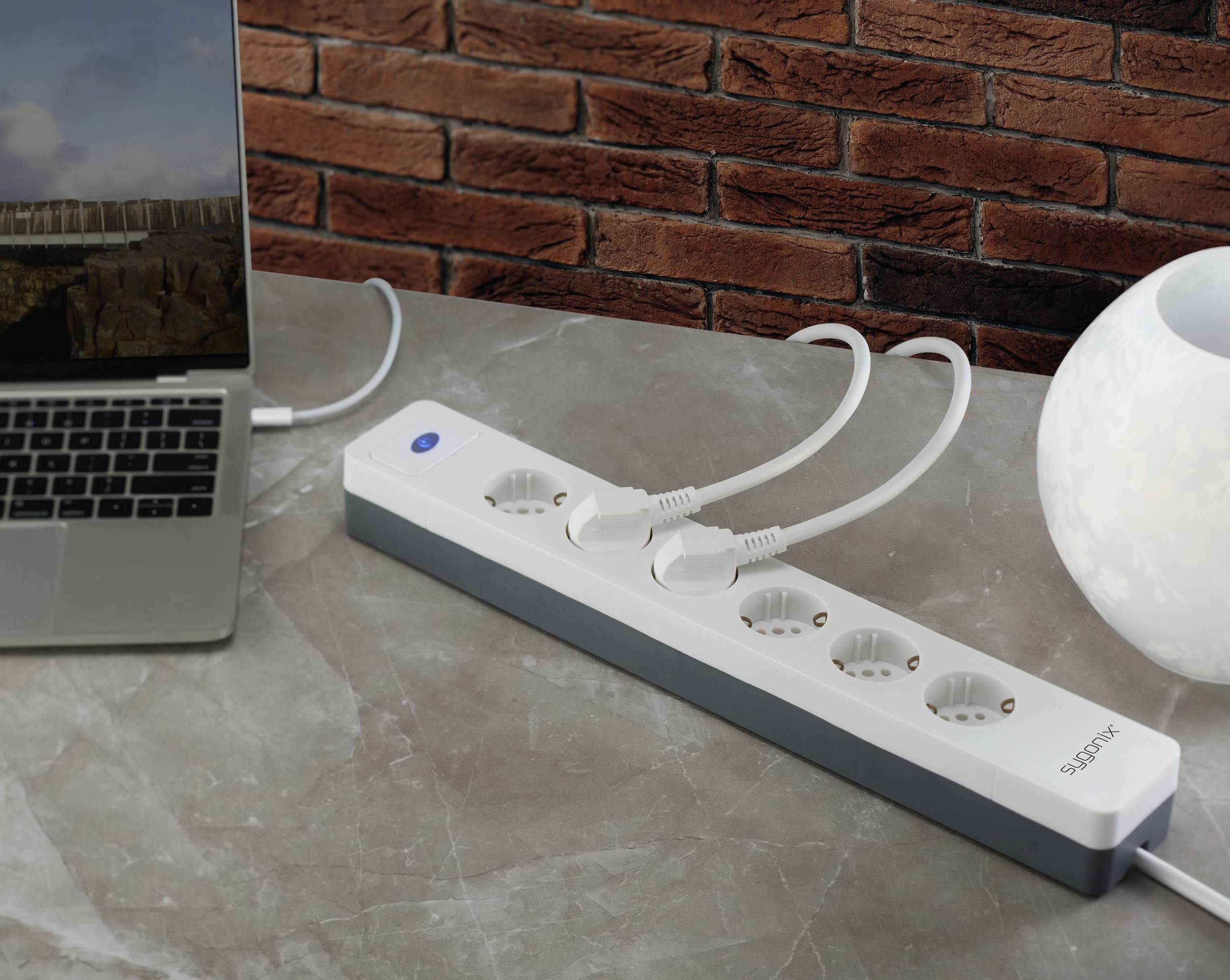 Eine Mehrfachsteckdose mit zwei eingesteckten Kabeln liegt auf einem grauen Tisch neben einem Laptop und einer weißen Lampe.