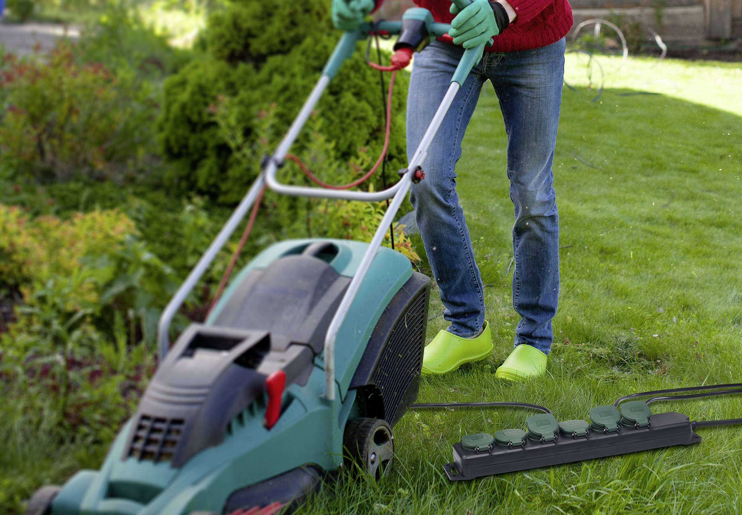 Eine Person mäht den Rasen mit einem Rasenmäher. Eine Steckdosenleiste liegt im Gras.