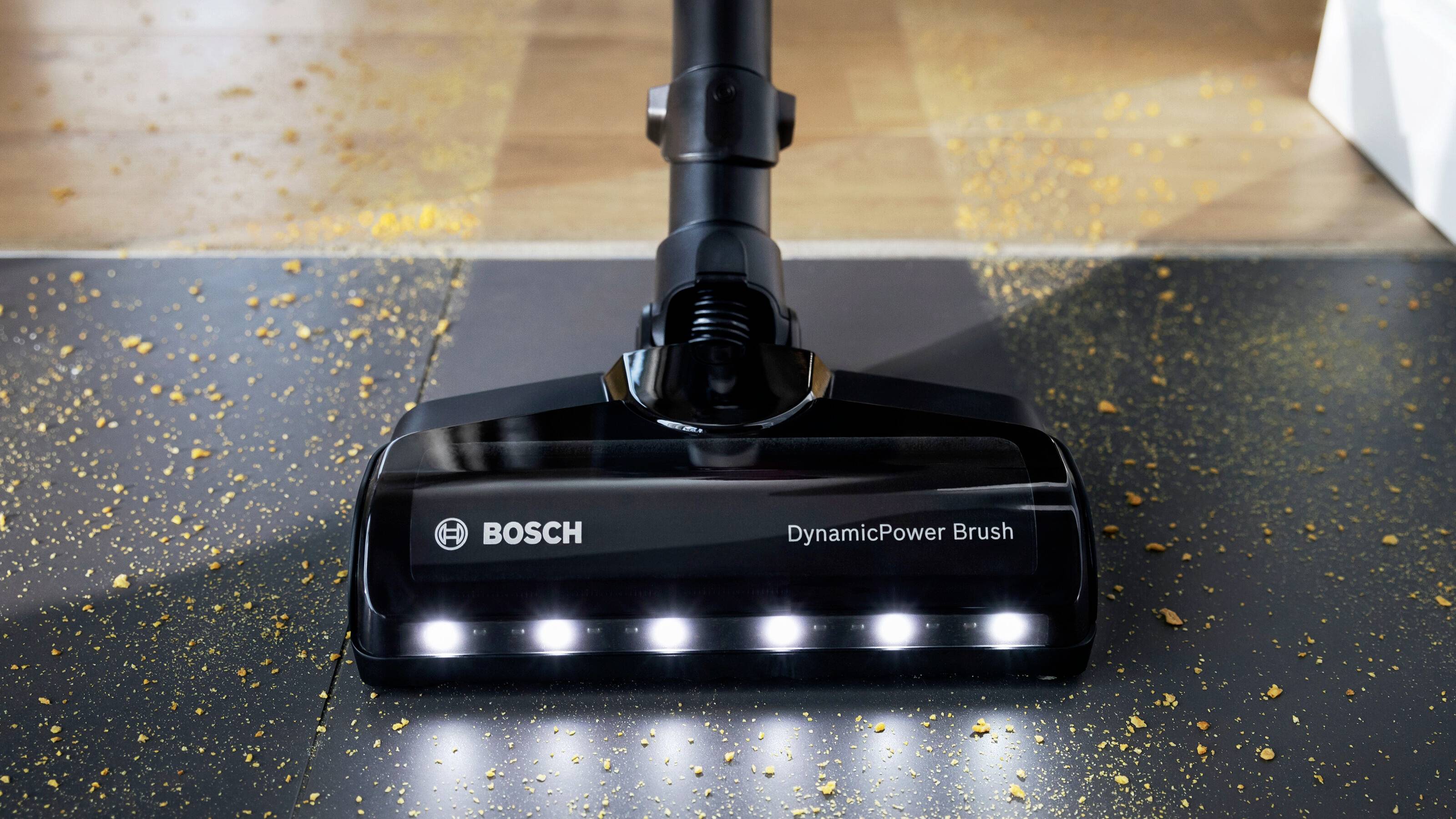 Ein Staubsauger von Bosch, Modell 'DynamicPower Brush', saugt Krümel von einem Boden auf. Beleuchtete Bürste zeigt Reinigungsstärke.