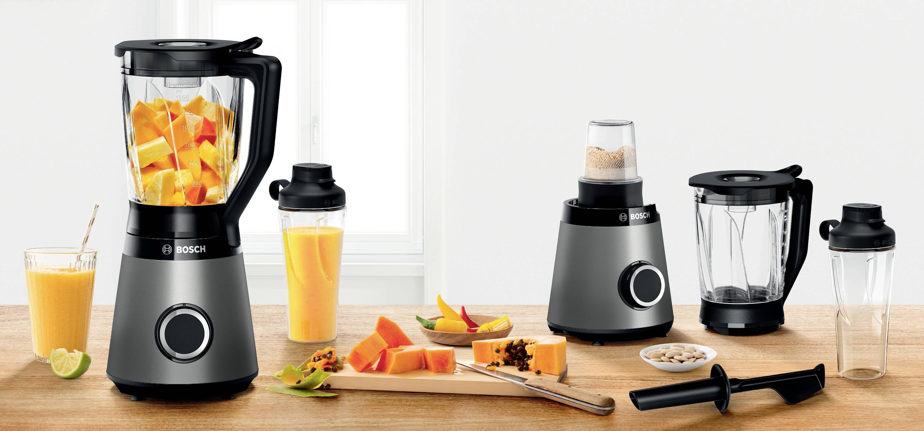 Mehrere Küchengeräte auf einem Tisch: Ein Standmixer mit geschnittenen Mango-Stücken, ein Mixbehälter, ein Zerkleinerer und Zubehör.