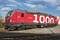 Eine rote Lokomotive mit der Nummer '1000' und der Aufschrift 'VECTRON' steht auf einem Bahngleis. Im Hintergrund ist blauer Himmel.
