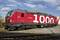 Roter Lokomotiv mit der Aufschrift '1000', im Freien neben Bahngleisen. Eine herbstliche Umgebung mit blauem Himmel und Wolken.