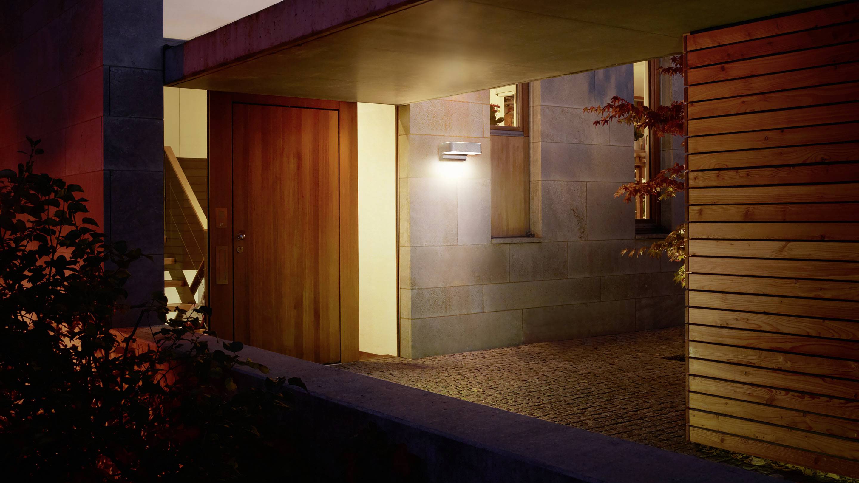 Eingangsbereich eines Hauses bei Dämmerung mit weißem Außenlicht an der Wand, Holztür und moderner Architektur umgeben von Pflanzen.