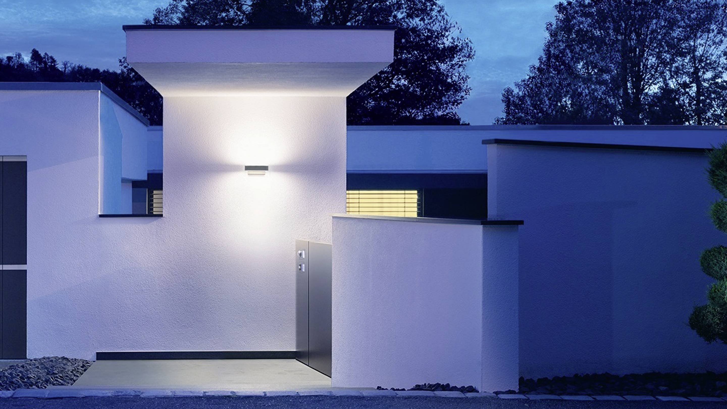 Ein modernes Gebäude bei Dämmerung mit beleuchtetem Eingang. Das Licht wirft Schatten auf die minimalistischen, weißen Wände.