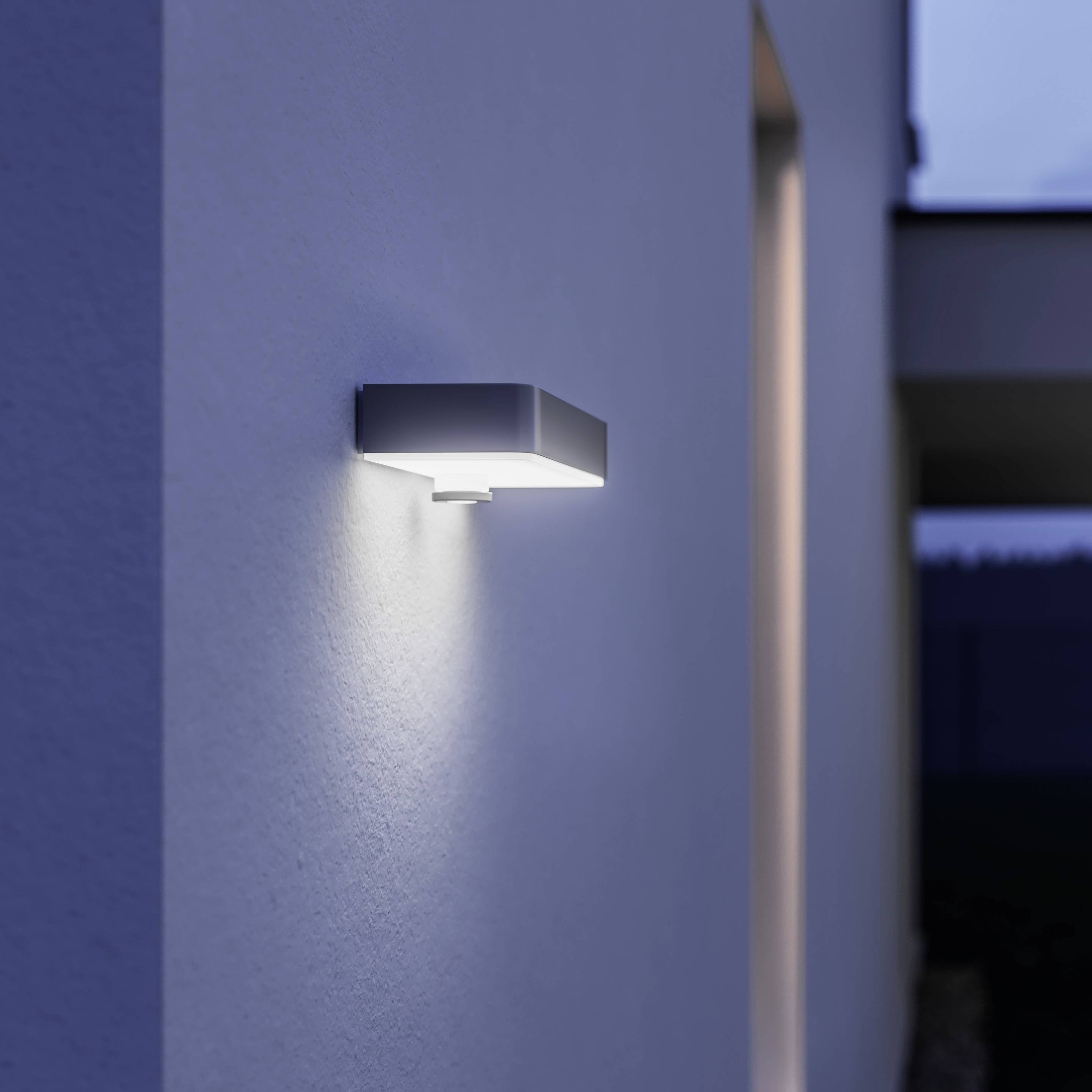 Eine moderne Außenlampe an einer Hauswand leuchtet in der Dämmerung. Sie spendet Licht auf die Fassade und den Gehweg.