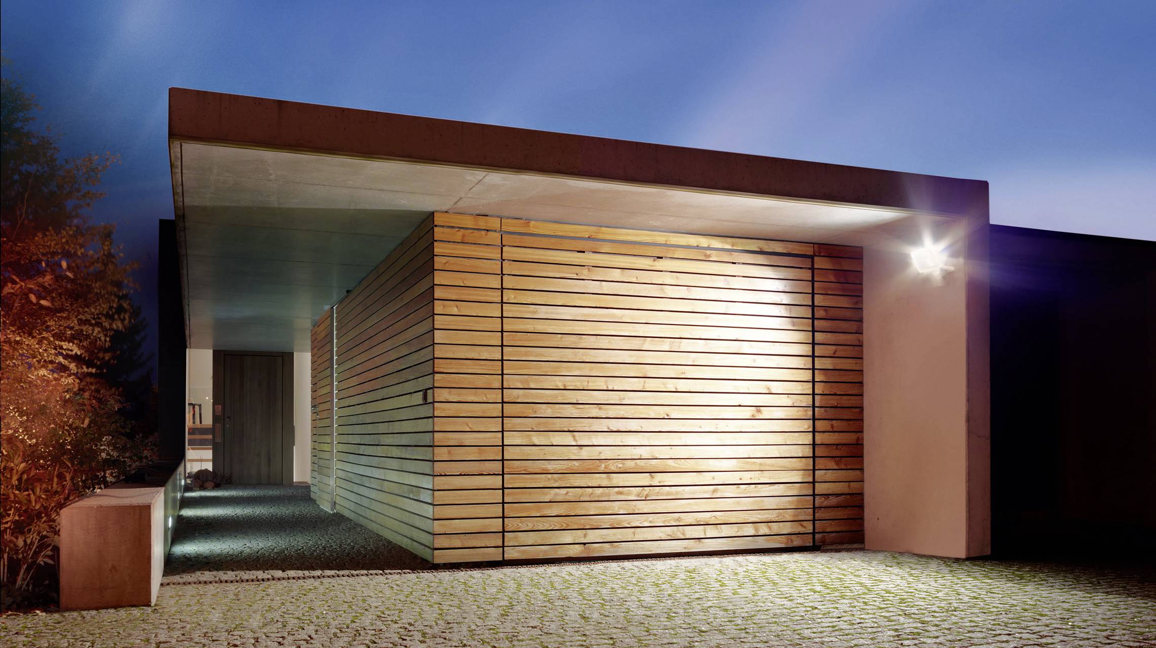 Modernes Haus mit Holzfassade und Flachdach, beleuchtet bei Dämmerung. Links ist ein beleuchteter Weg, der zur Eingangstür führt.