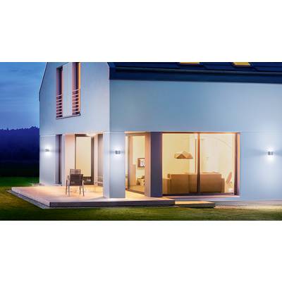 Moderne Hausfassade bei Abenddämmerung mit beleuchtetem Wohnzimmer und Terrasse mit zwei Stühlen, umgeben von einem Rasen.