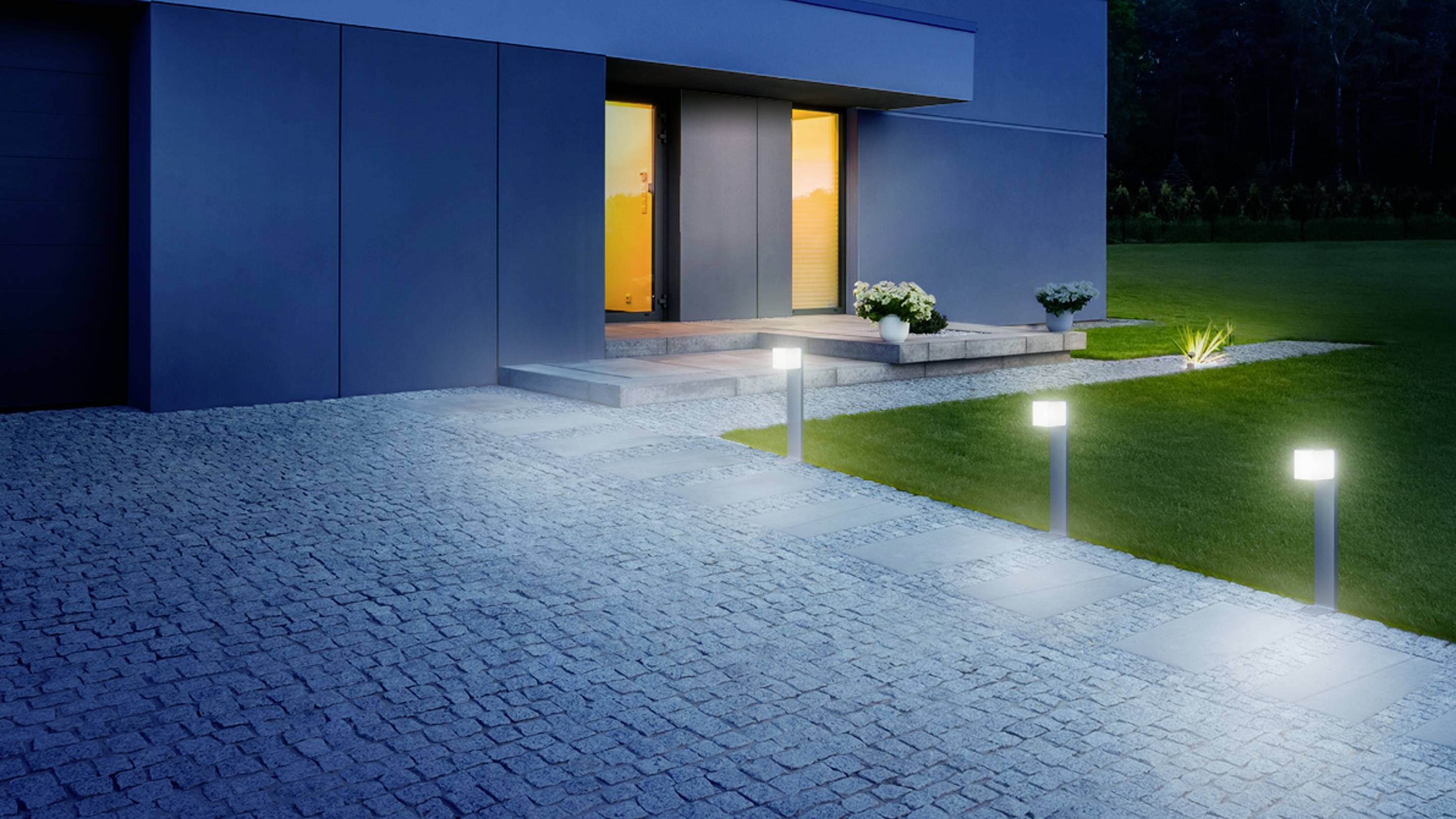 Eingangsbereich eines modernen Hauses bei Dämmerung mit beleuchtetem Gartenweg und der Tür, die in warmem Licht leuchtet.