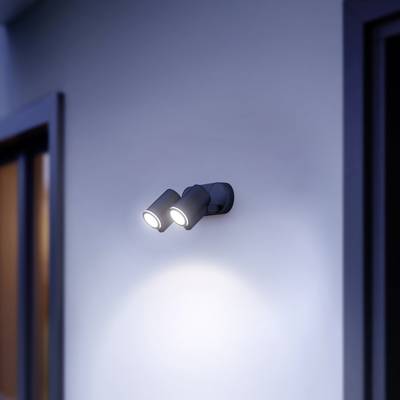 Eine Außenwand mit einer modernen, zweistrahligen Wandlampe beleuchtet den Bereich darunter. Auf der linken Seite ist ein Fenster.