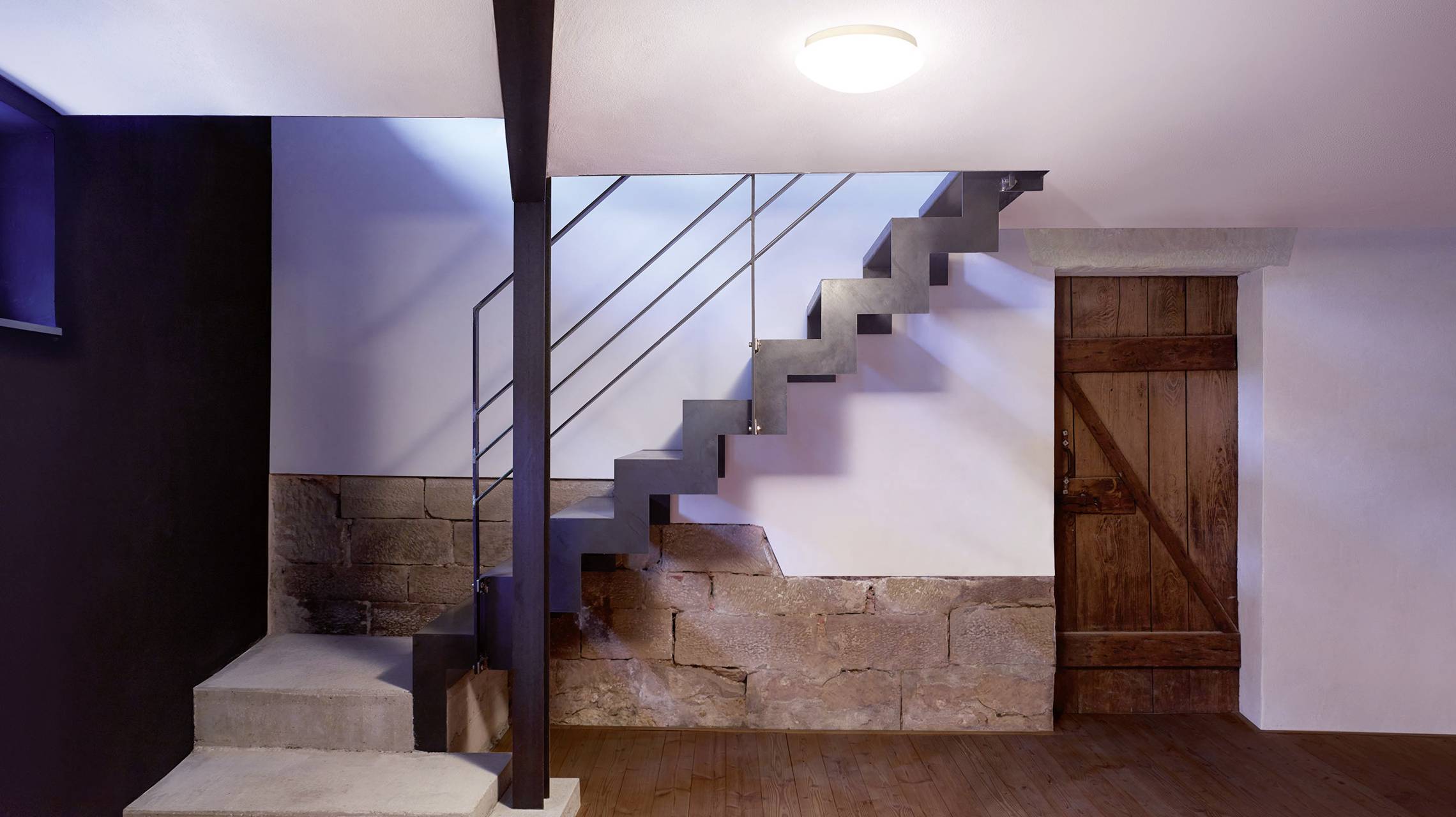 Ein moderner Kellerraum mit Metalltreppe, gemauerter Wand und Holztür. Das Licht von der Decke erhellt den Raum.