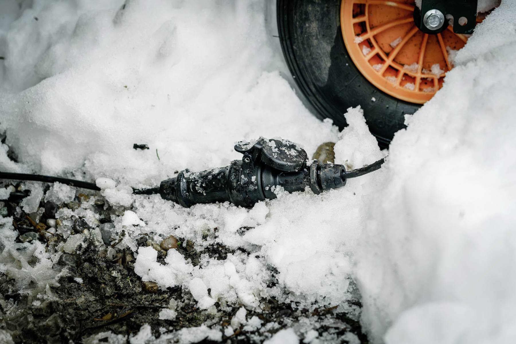 Ein schwarzer Schlauch mit einem Ventil liegt auf Schnee und Matsch neben einem großen Rad mit orangener Felge.
